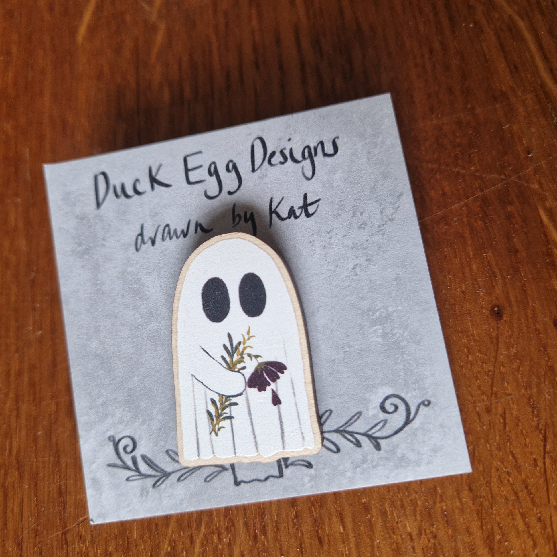A white ghost pin badge sits on a grey backing card with the words 'Duck Egg Designs drawn by Kat' across the top. The ghost is holding a drooping red flower and some leaves. Behind the backing card is a wooden background.