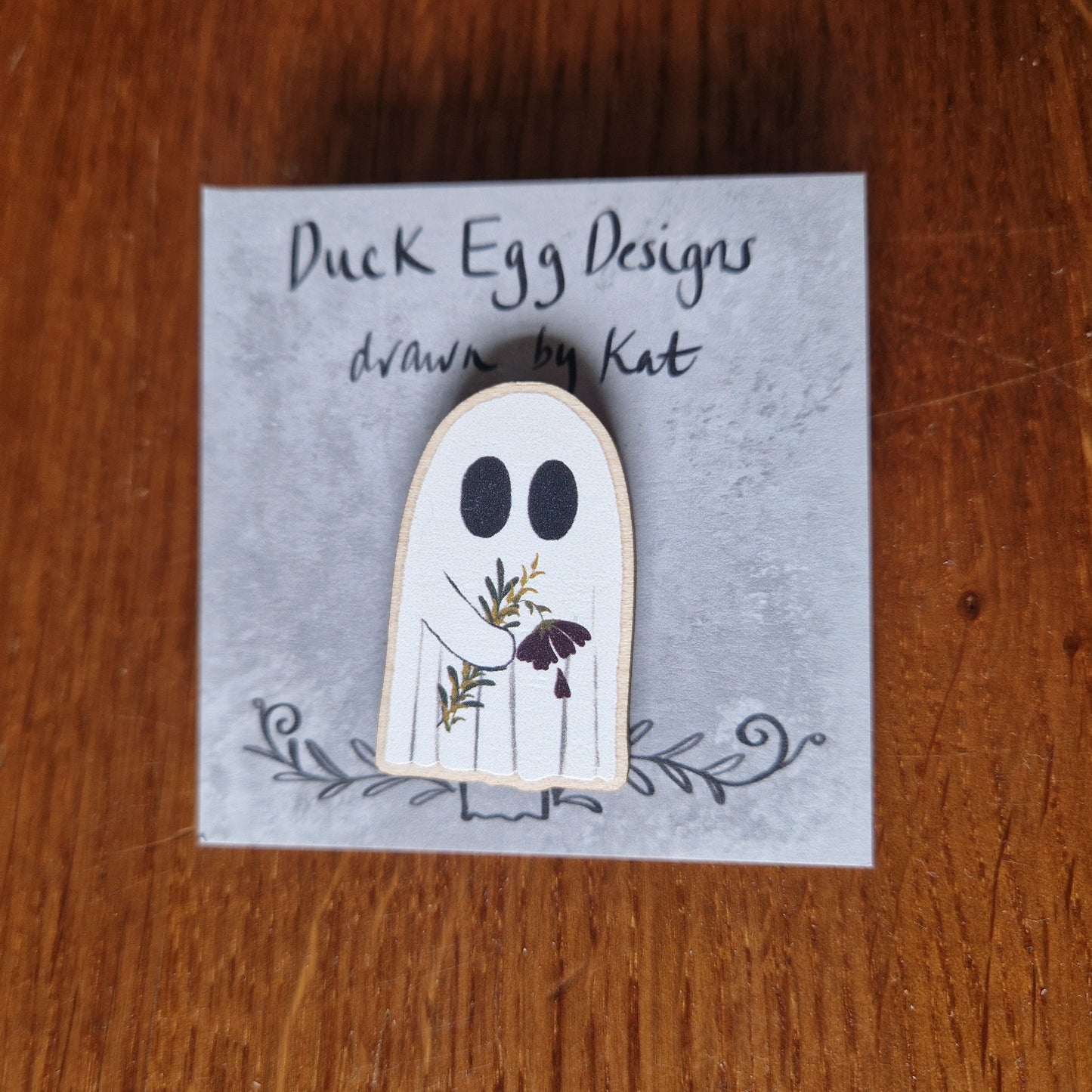 A white ghost pin badge sits on a grey backing card with the words 'Duck Egg Designs drawn by Kat' across the top. The ghost is holding a drooping red flower and some leaves. Behind the backing card is a wooden background.
