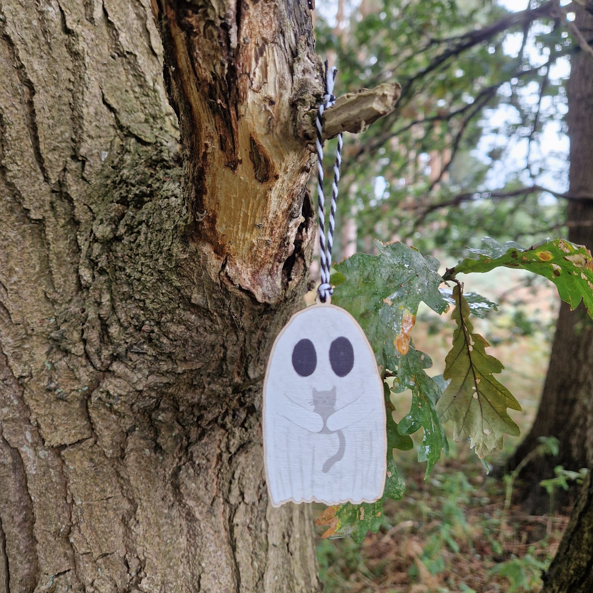 A white ghost holding a grey cat hangs by a white and black string from a green leafed tree.