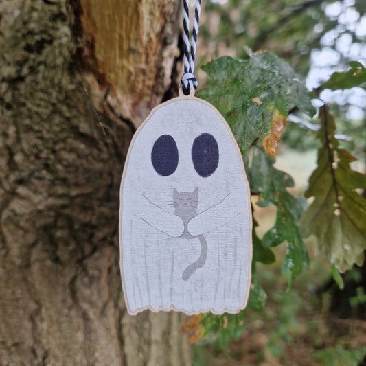 A white ghost holding a grey cat hangs by a white and black string from a green leafed tree.