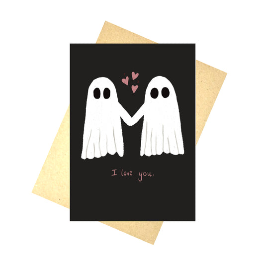 A black card with two ghosts holding hands with pink hearts in the air between them. Under the ghosts are the words 'I love you.' in pink handwriting. Behind the card is a brown envelope and a white background.