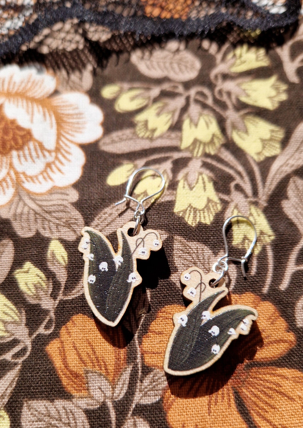 A pair of lily of the valley ghost flower earrings with sterling silver hooks lie on a retro floral fabric with a black lace fabric visible at the top of the image.