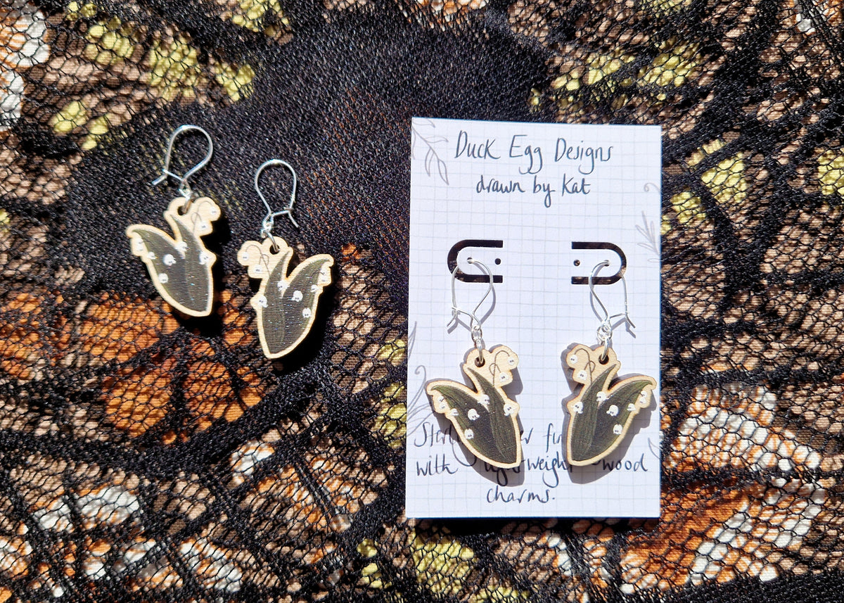 Two pair of ghost lily of the valley earrings sit next to each other on top of a black lace and floral fabric background.  The pair on the right is on a white backing card with the words 'Duck Egg Designs drawn by Kat' across the top.