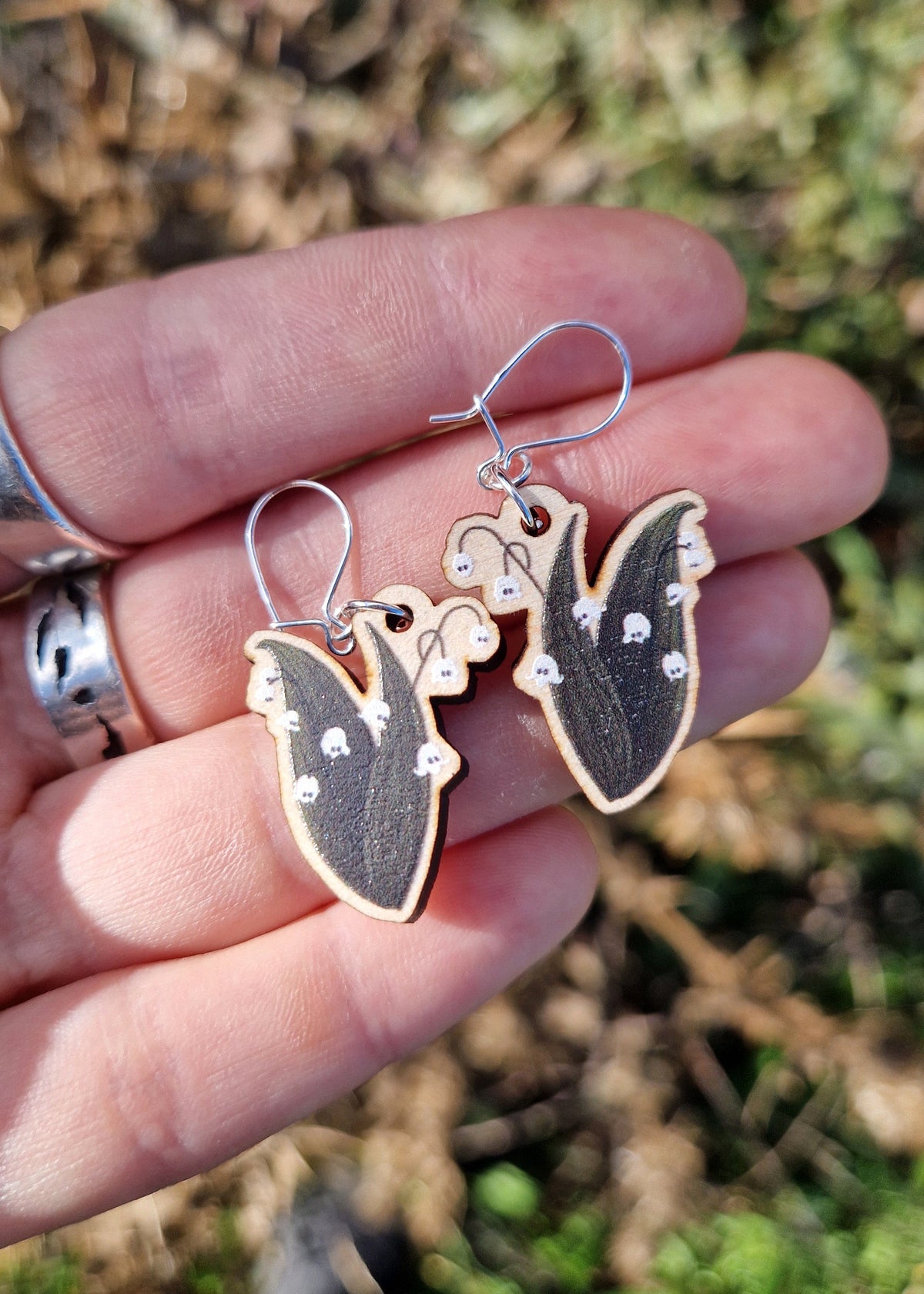 A pair of ghost lily of the valley earrings showing the dark green, white and black charms with the sterling silver jump rings and earring hooks. The earrings sit on a white hand wearing silver rings above an out of focus garden.