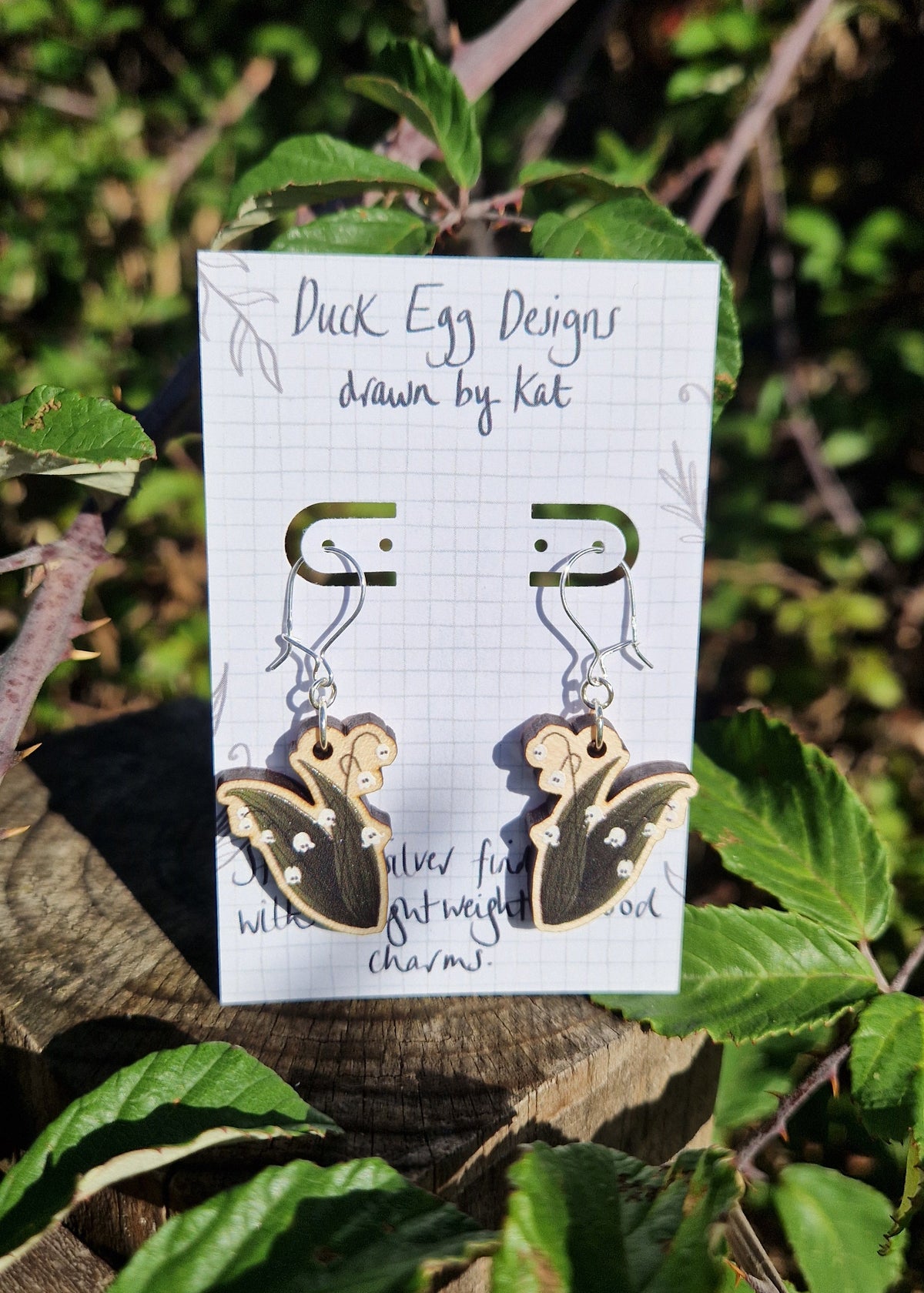 A pair of ghost lily of the valley earrings sit on a backing card on a wooden post surrounded by bramble leaves. 