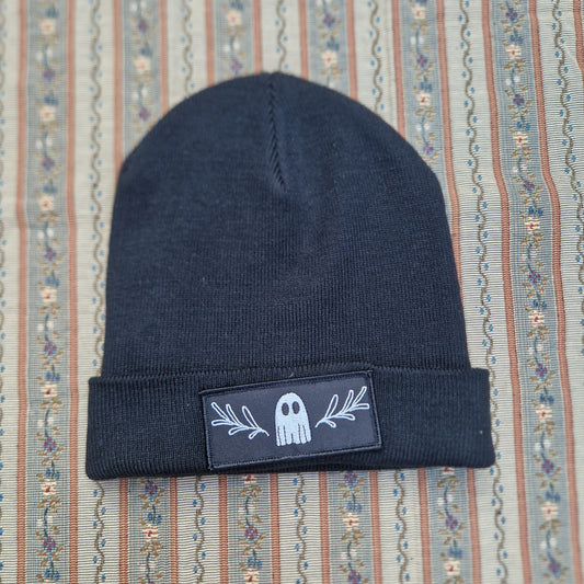A black beanie with a folded up bottom with a rectangular black patch with a ghost and symmetrical leafy vine design. Behind the beanie is a striped vintage fabric.