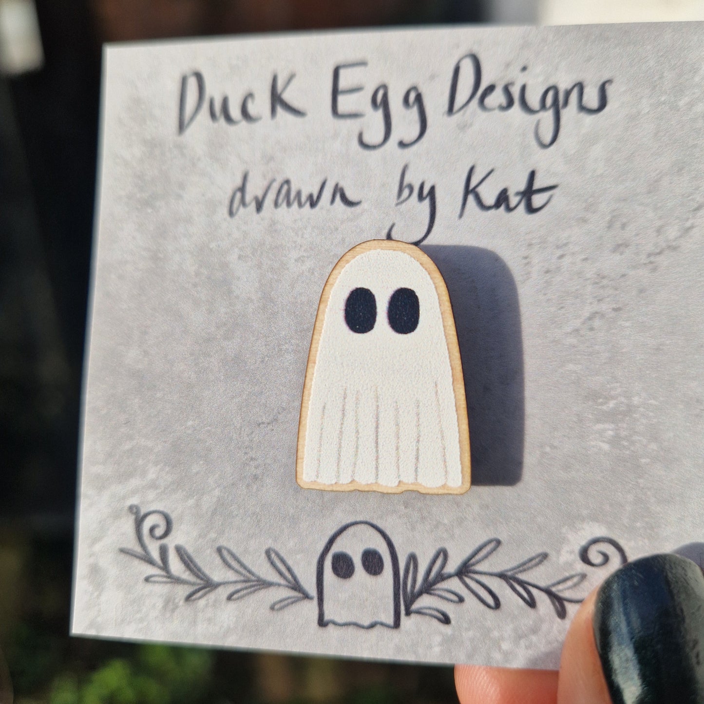 A closer look at the wooden ghost pin badge, showing the texture of the pin. The white ghost pin sits on a mottled grey square backing card with the words 'Duck Egg Designs drawn by Kat'.