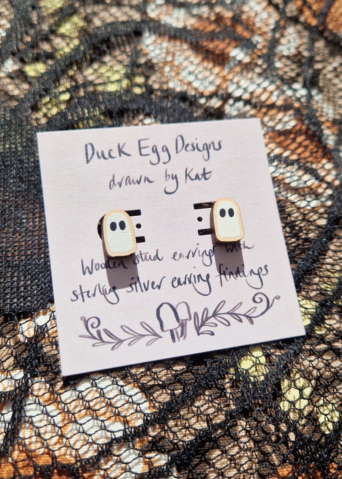 A pair of white ghost studs sit on a light purple backing card that has the words 'Duck Egg Designs drawn by Kat' in black writing. Behind the backing card is a black lace fabric over a floral fabric.