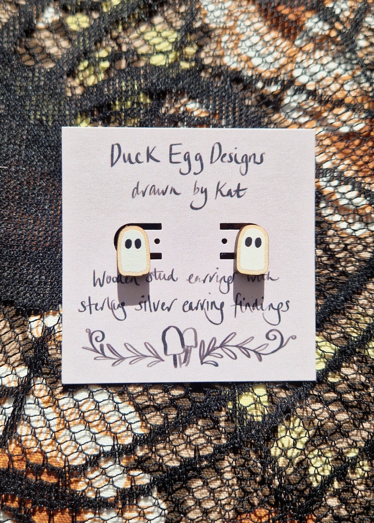 A pair of white ghost studs sit on a light purple backing card that has the words 'Duck Egg Designs drawn by Kat' in black writing. Behind the backing card is a black lace fabric over a floral fabric.