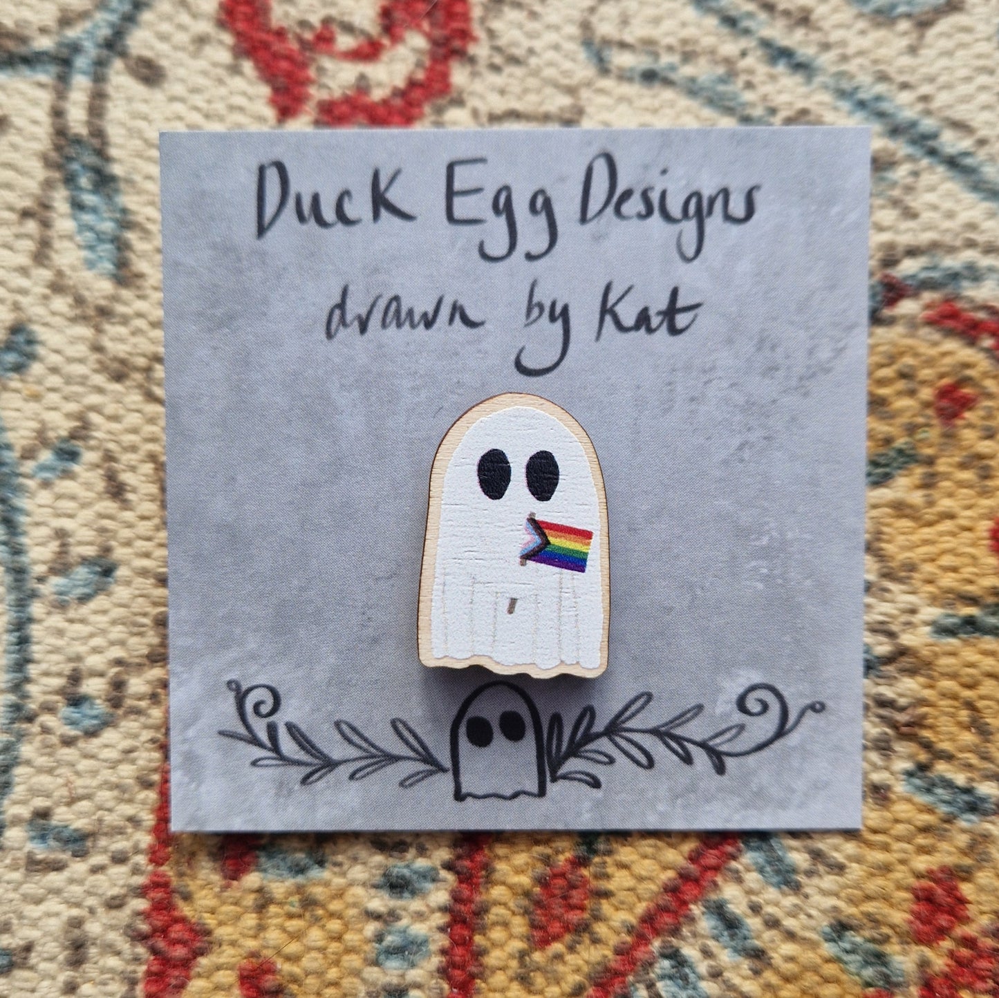 A pin of a white ghost holding the progress LGBTQ+ flag on a grey backing card with the black words 'Duck Egg Designs drawn by Kat'. Behind the backing card is a textured fabric background.