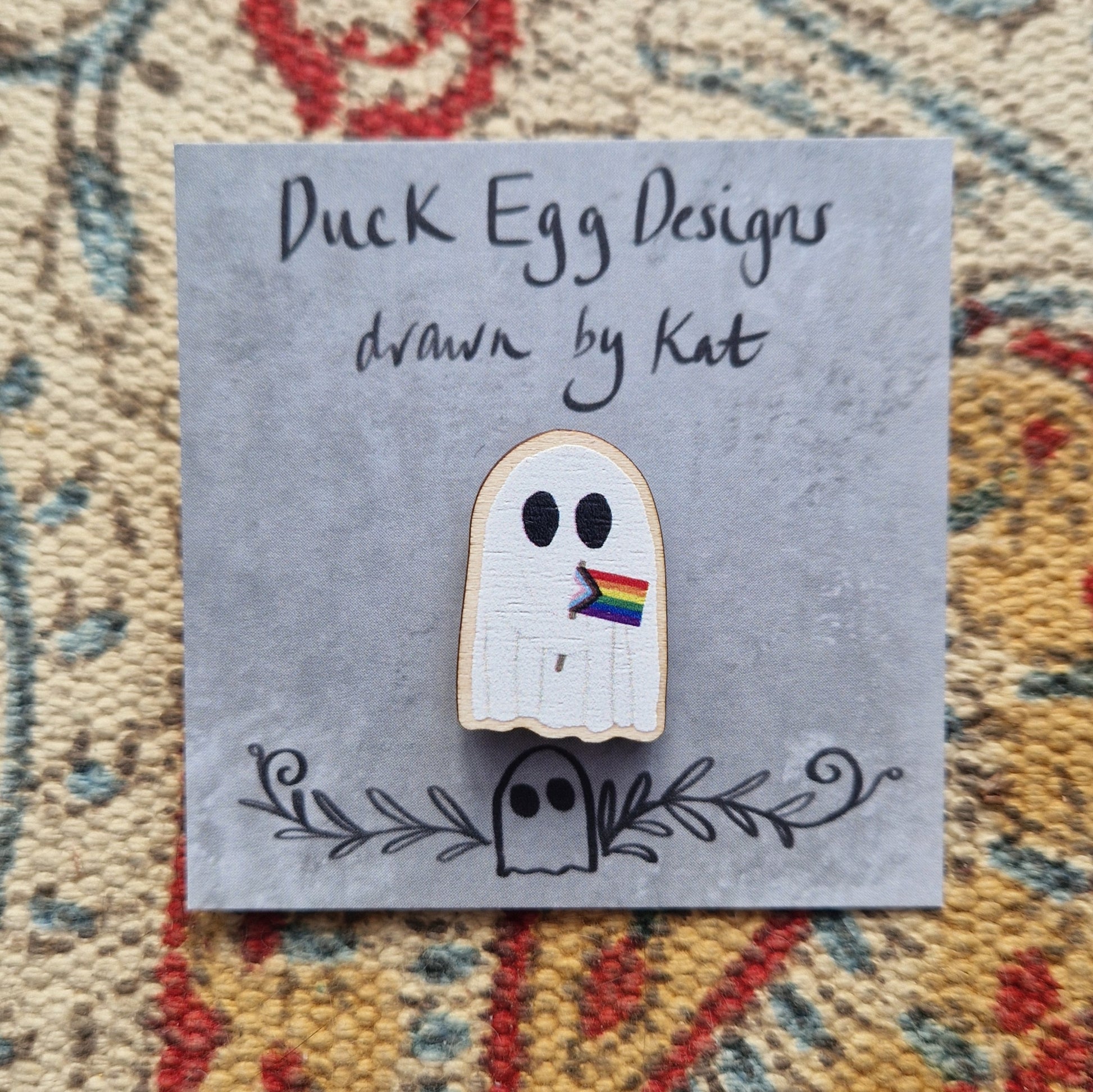 A pin of a white ghost holding the progress LGBTQ+ flag on a grey backing card with the black words 'Duck Egg Designs drawn by Kat'. Behind the backing card is a textured fabric background.