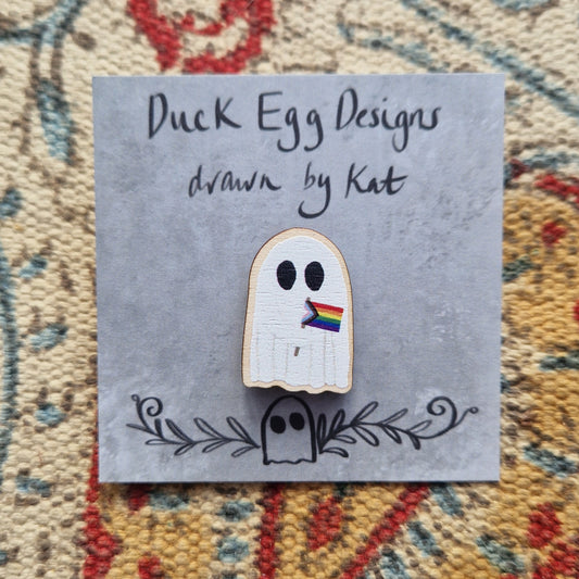 A pin of a white ghost holding the progress LGBTQ+ flag on a grey backing card with the black words 'Duck Egg Designs drawn by Kat'. Behind the backing card is a textured fabric background.