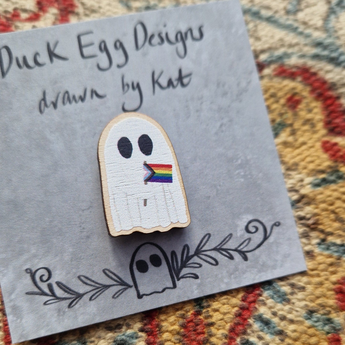 A pin of a white ghost holding the progress LGBTQ+ flag on a grey backing card with the black words 'Duck Egg Designs drawn by Kat'. Behind the backing card is a textured fabric background.