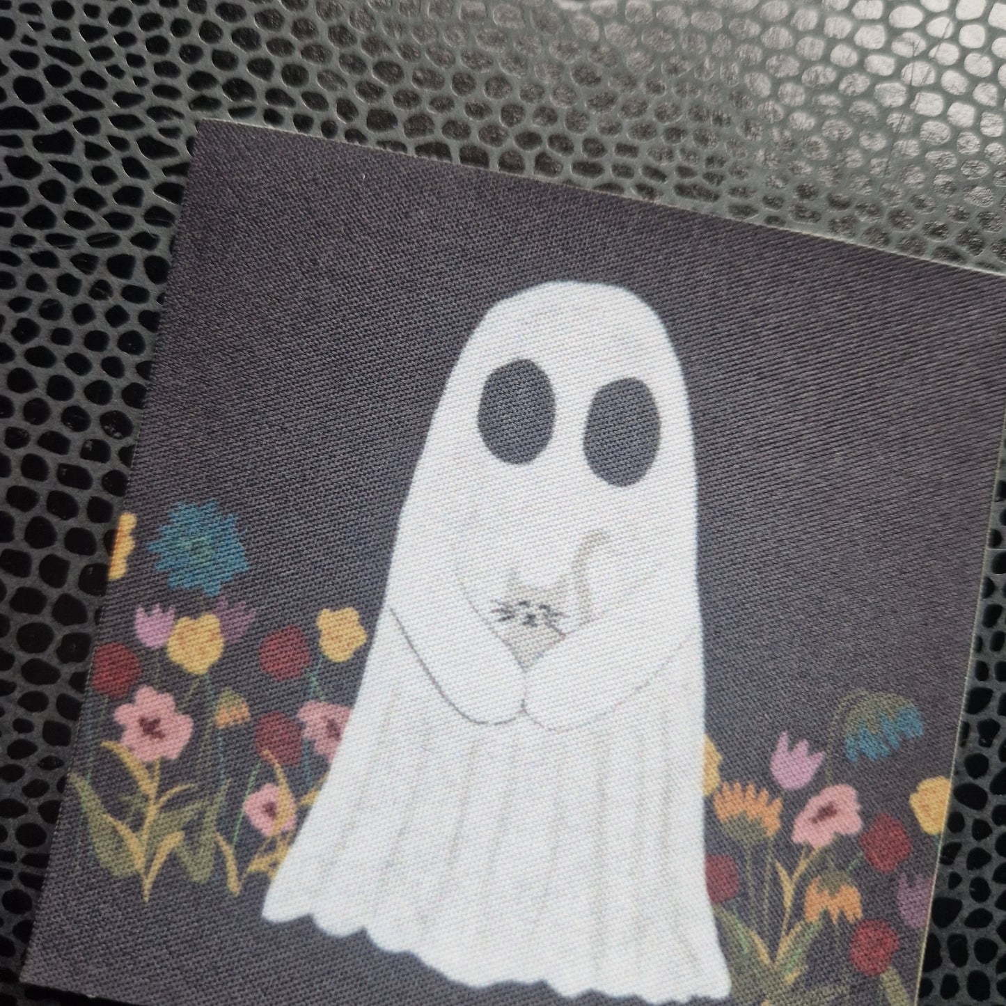 A closer look at the ghost patch showing the texture of the fabric. A ghost carrying a cat surrounded by red, pink, yellow and blue flowers on a square black patch. Behind the patch is a black and grey printed snakeskin patterned bookcover.