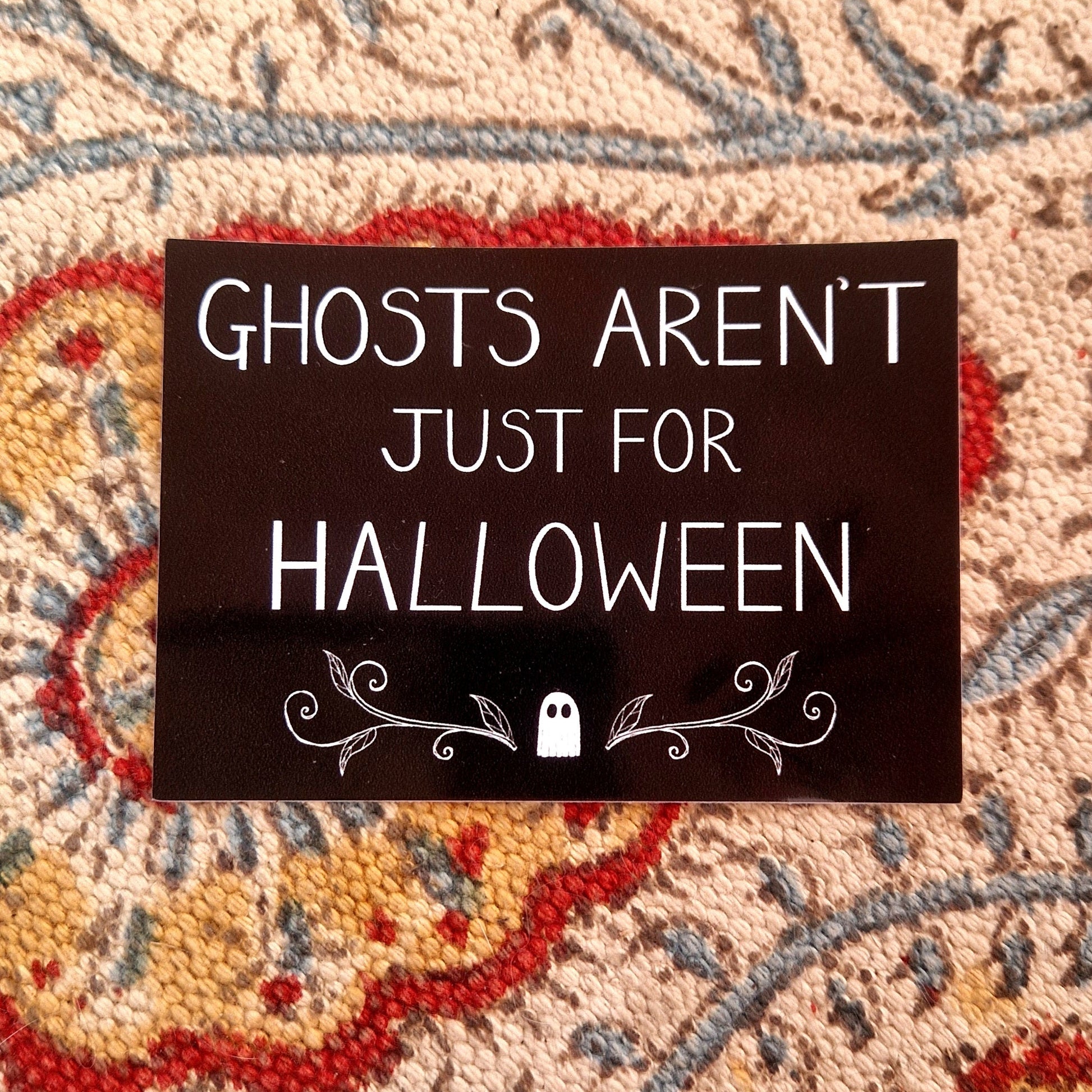 A black rectangular sticker featuring the words 'GHOSTS AREN'T JUST FOR HALLOWEEN" in white handwriting above a ghost and symmetrical vines design. Behind the sticker is a patterned fabric background.