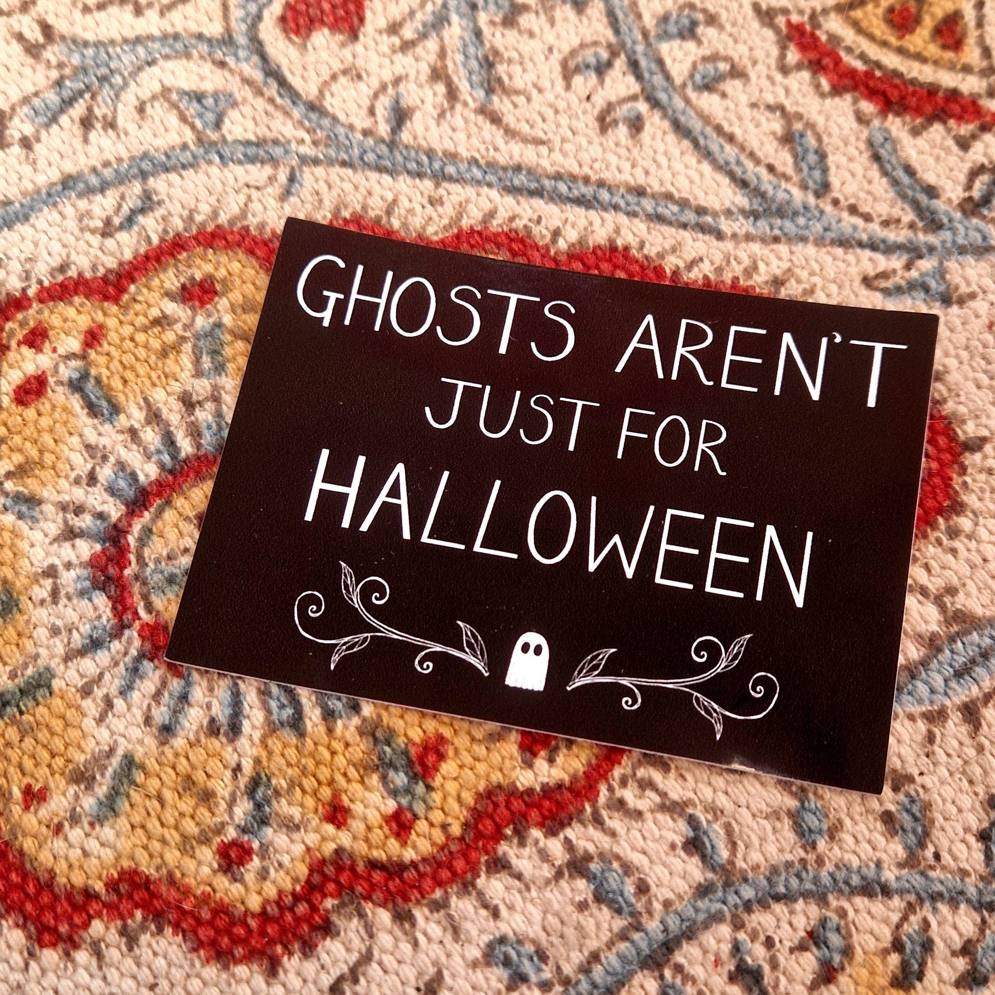 A black rectangular sticker featuring the words 'GHOSTS AREN'T JUST FOR HALLOWEEN" in white handwriting above a ghost and symmetrical vines design. Behind the sticker is a patterned fabric background.