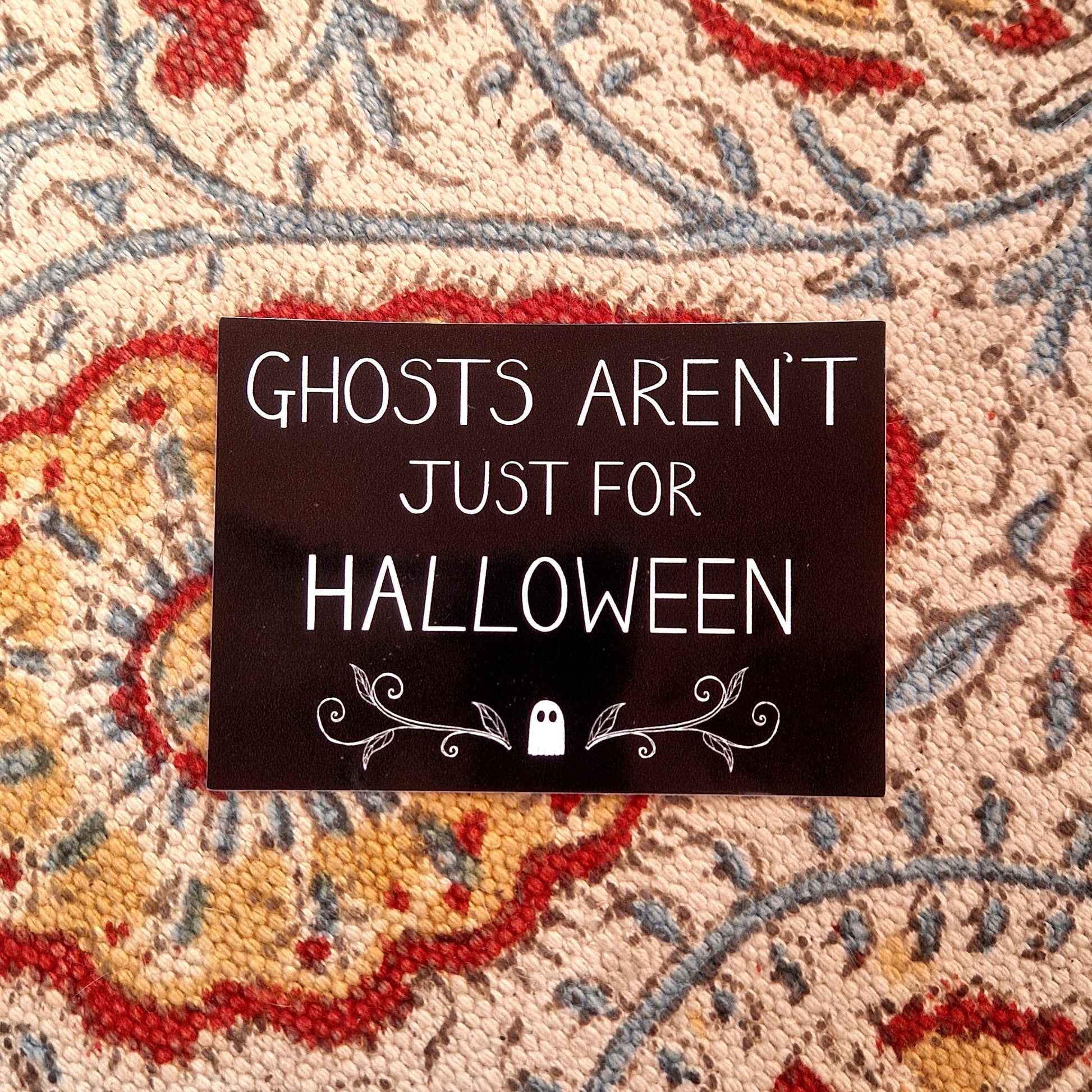 A black rectangular sticker featuring the words 'GHOSTS AREN'T JUST FOR HALLOWEEN" in white handwriting above a ghost and symmetrical vines design. Behind the sticker is a patterned fabric background.
