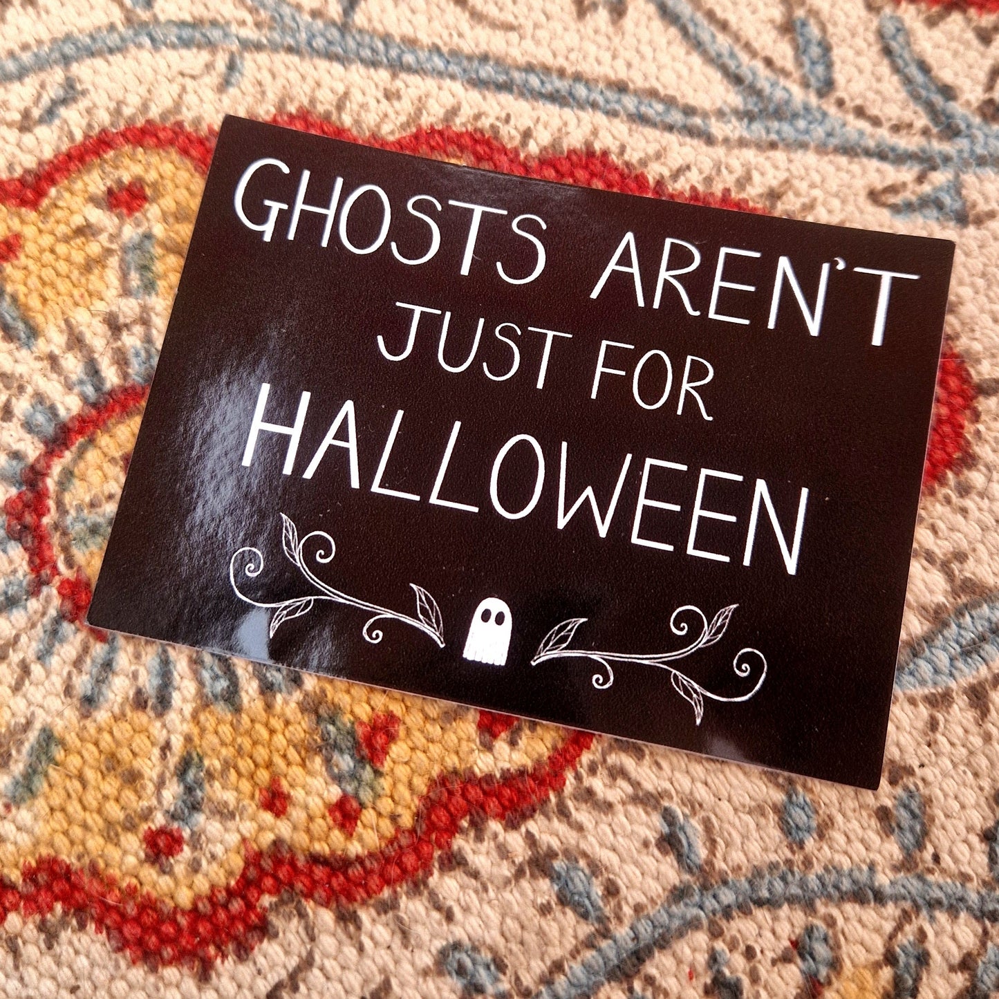 A black rectangular sticker featuring the words 'GHOSTS AREN'T JUST FOR HALLOWEEN" in white handwriting above a ghost and symmetrical vines design. Behind the sticker is a patterned fabric background.