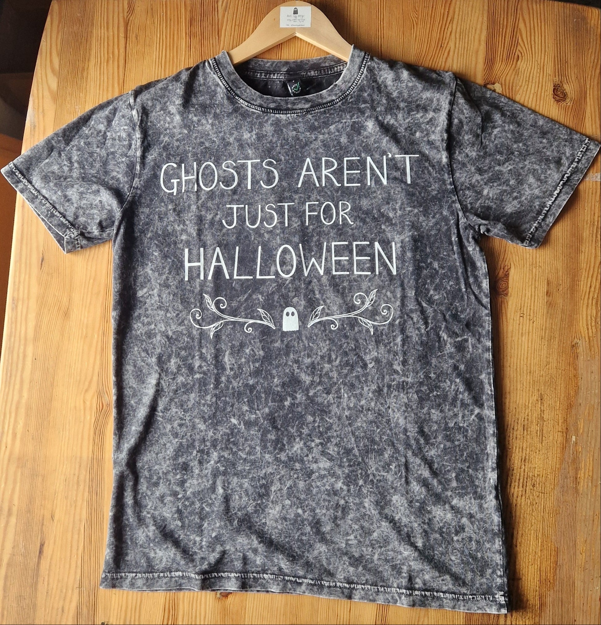 A mottled grey t-shirt with the words 'Ghosts Aren't Just For Halloween' in white writing above a ghost and leafy tendril design. The t-shirt lies on a wooden table.