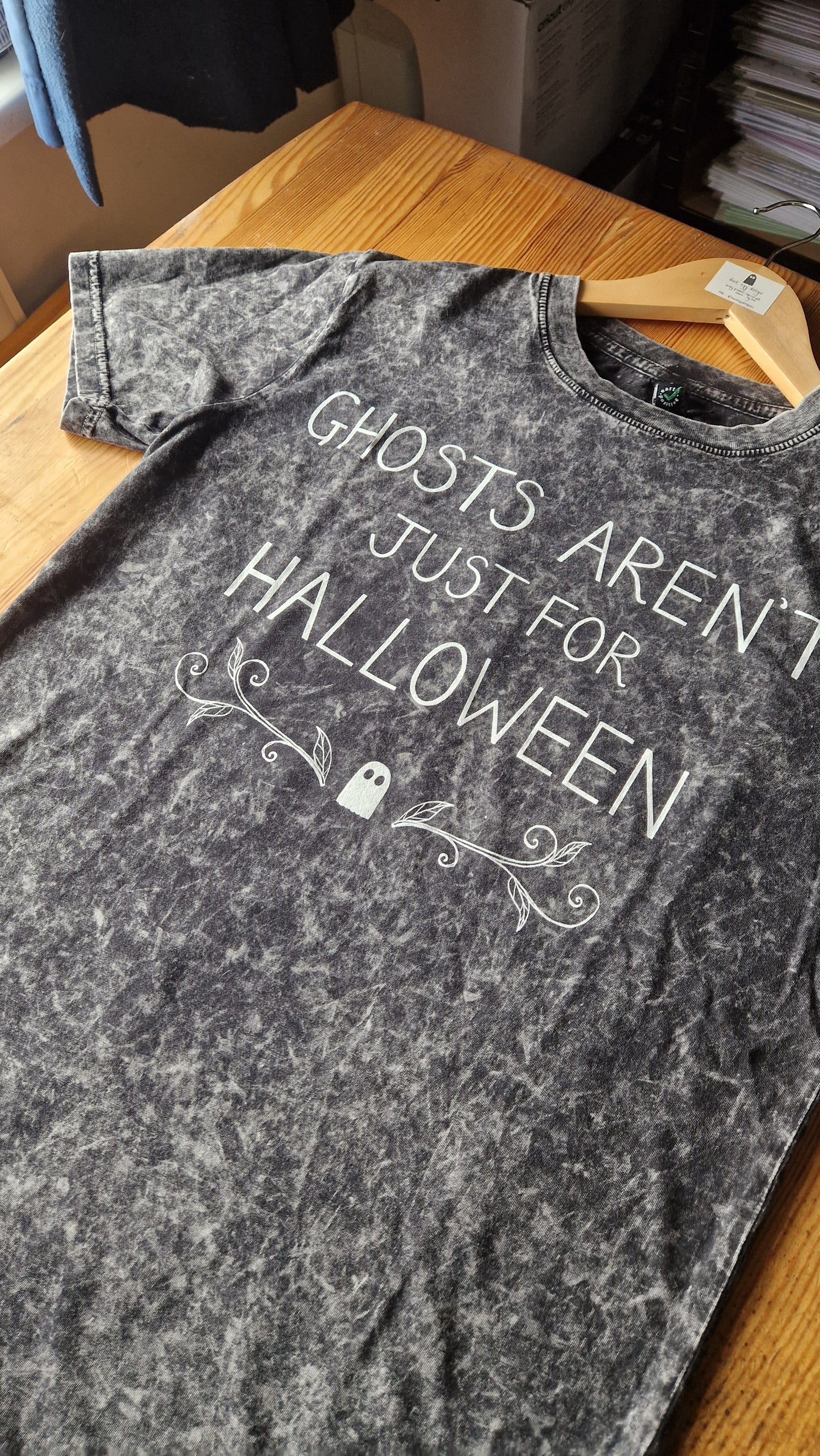 A mottled grey t-shirt lies on a wooden table. It's design features the words 'Ghosts Aren't Just For Halloween' in white handwriting above a small ghost with leafy tendrils on either side of it.