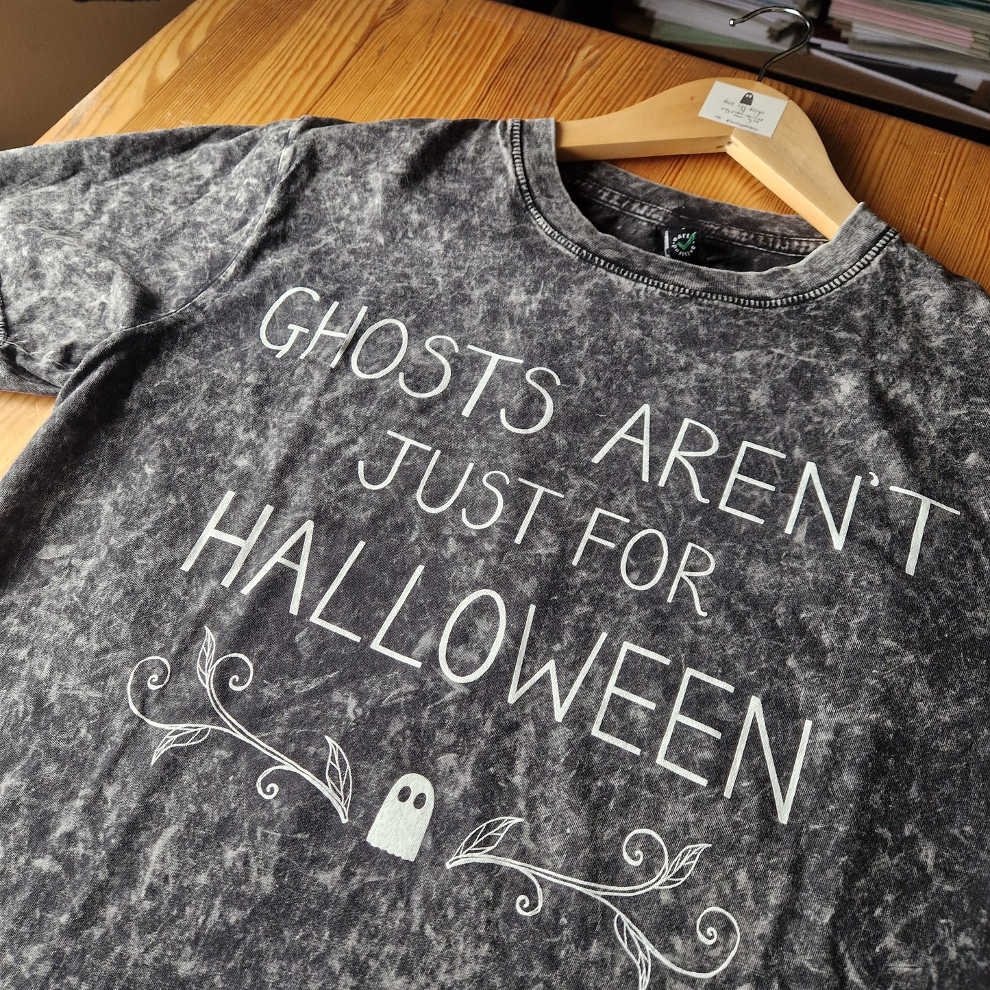 A close up of the mottled grey tshirt showing the acid wash of the fabric. A close up view of the handwritten text reading 'Ghosts Aren't Just For Halloween' as well as the ghost and leafy tendril design.