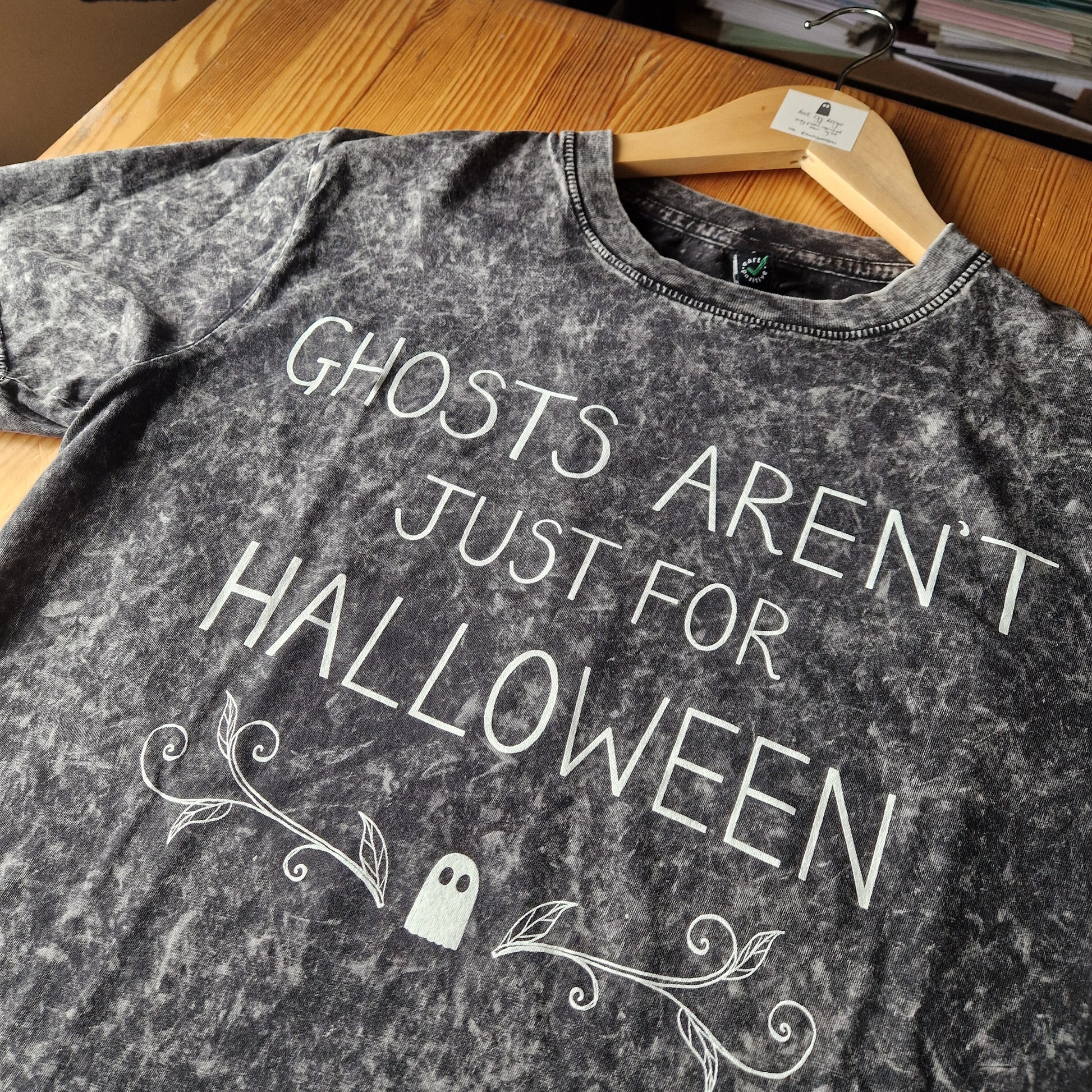 A close up of the mottled grey tshirt showing the acid wash of the fabric. A close up view of the handwritten text reading 'Ghosts Aren't Just For Halloween' as well as the ghost and leafy tendril design.