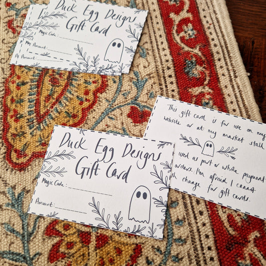 White gift cards with black designs sit on a textured patterned fabric background. The card says 'Duck Egg Designs Gift Card' across the front with space for a magic code and amount as well as a ghost and leafy tendrils growing in from the outside.