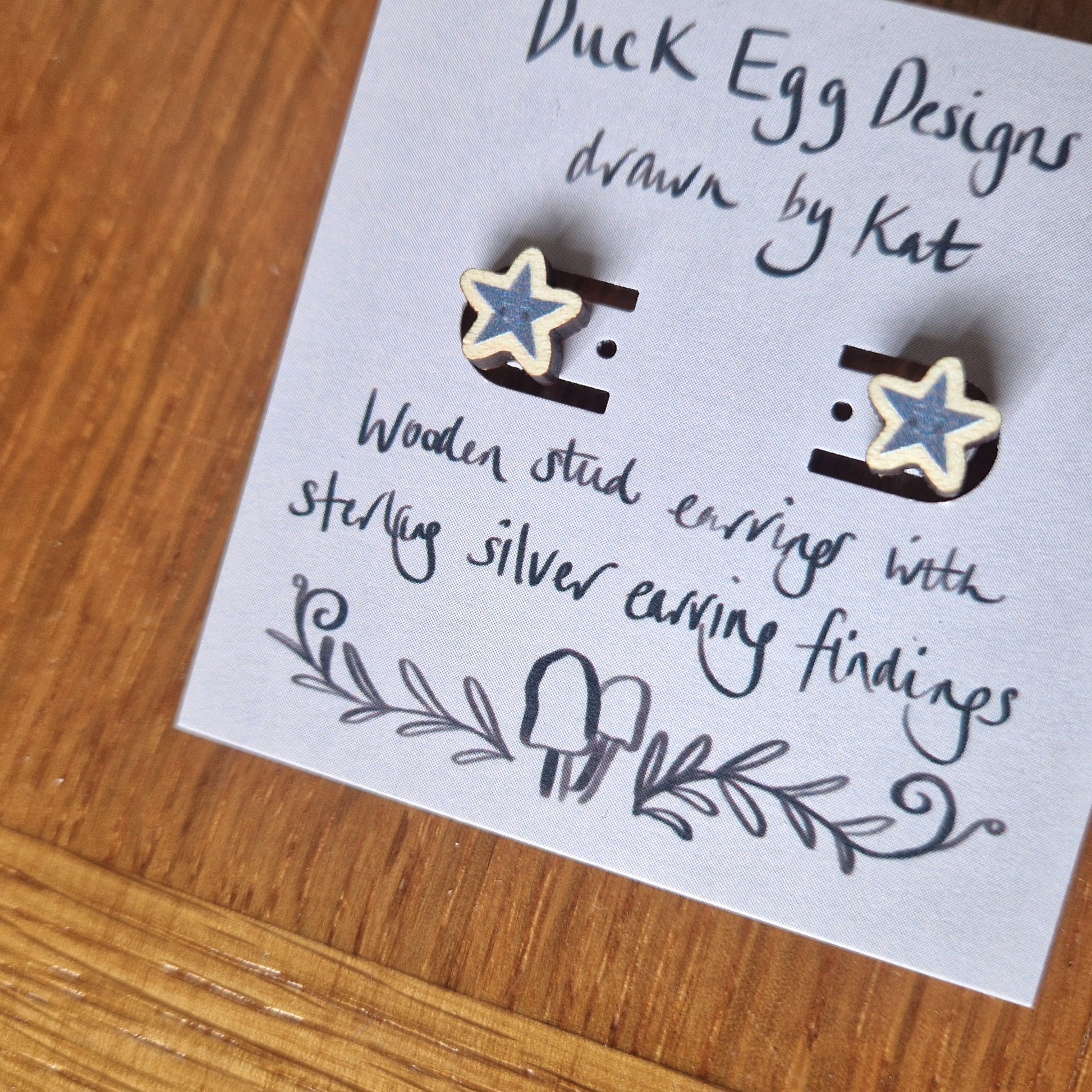 A close up of the pair of grey star earrings with small smiling faces sit on a light purple backing card with the words 'Duck Egg Designs drawn by Kat' across the top in black and 'Wooden stud earrings with sterling silver earring findings' across the bottom. Behind the backing card you can see a wooden background.
