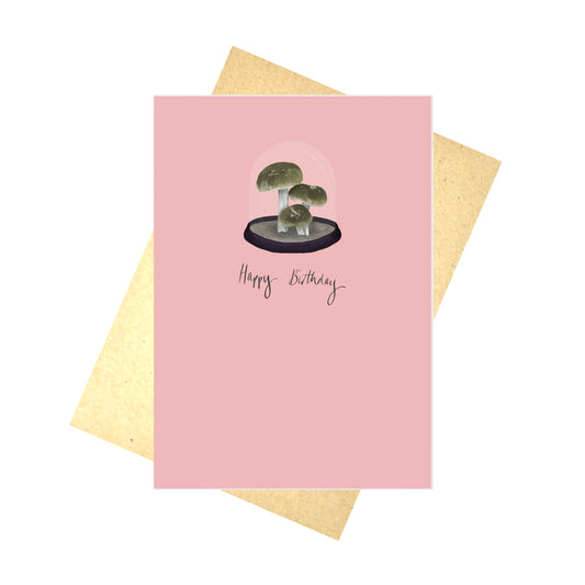A muted pink background with a fungi in a bell jar design and the words 'Happy Birthday' in deep purple handwriting. Behind the card is a brown envelope and a white background.