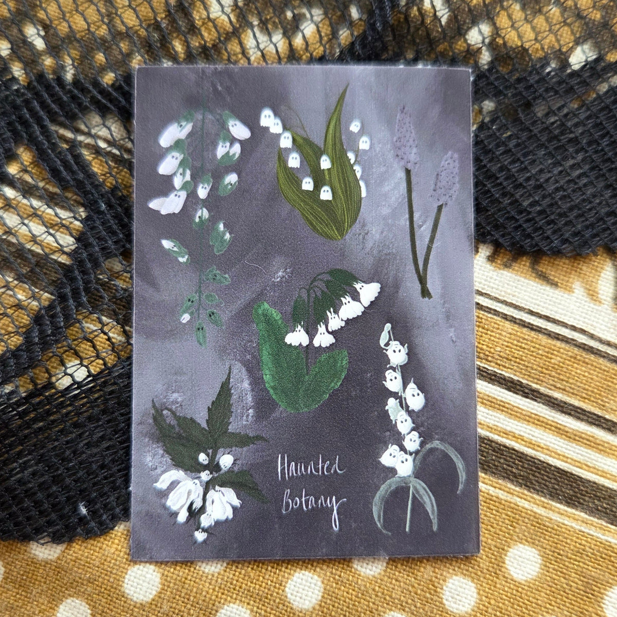 A purple and white rectangular sticker with six different haunted plant in whites and greens and the words 'Haunted Botany' at the bottom. Behind the sticker is a brown fabric with a lack lace overlay.