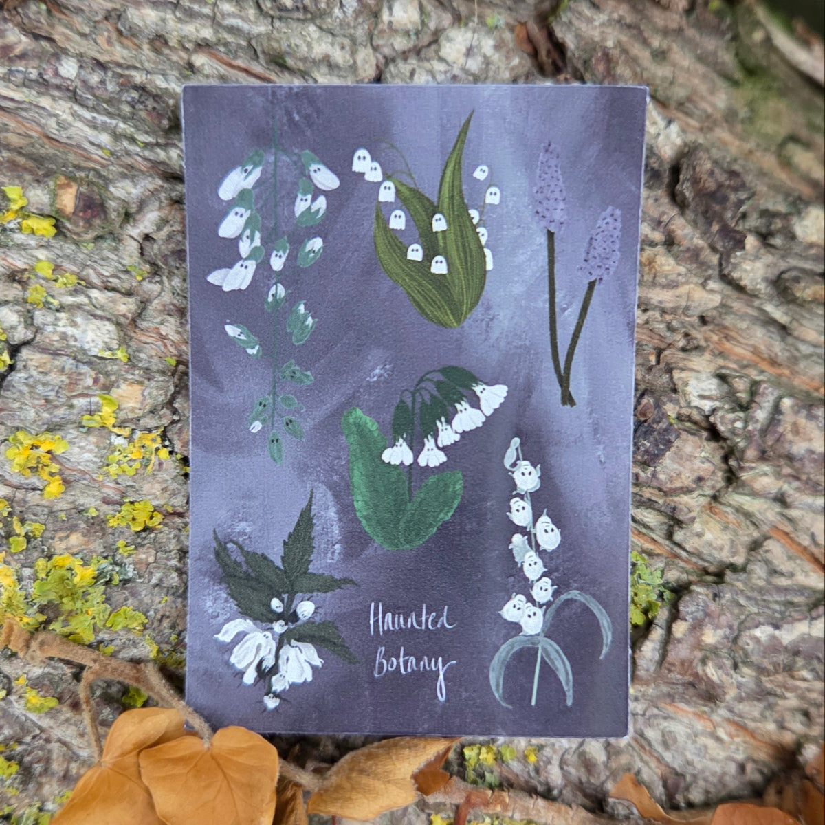 A purple and white rectangular sticker with six different haunted plant in whites and greens and the words 'Haunted Botany' at the bottom. Behind the sticker is the bark of a tree and some orange ivy leaves.
