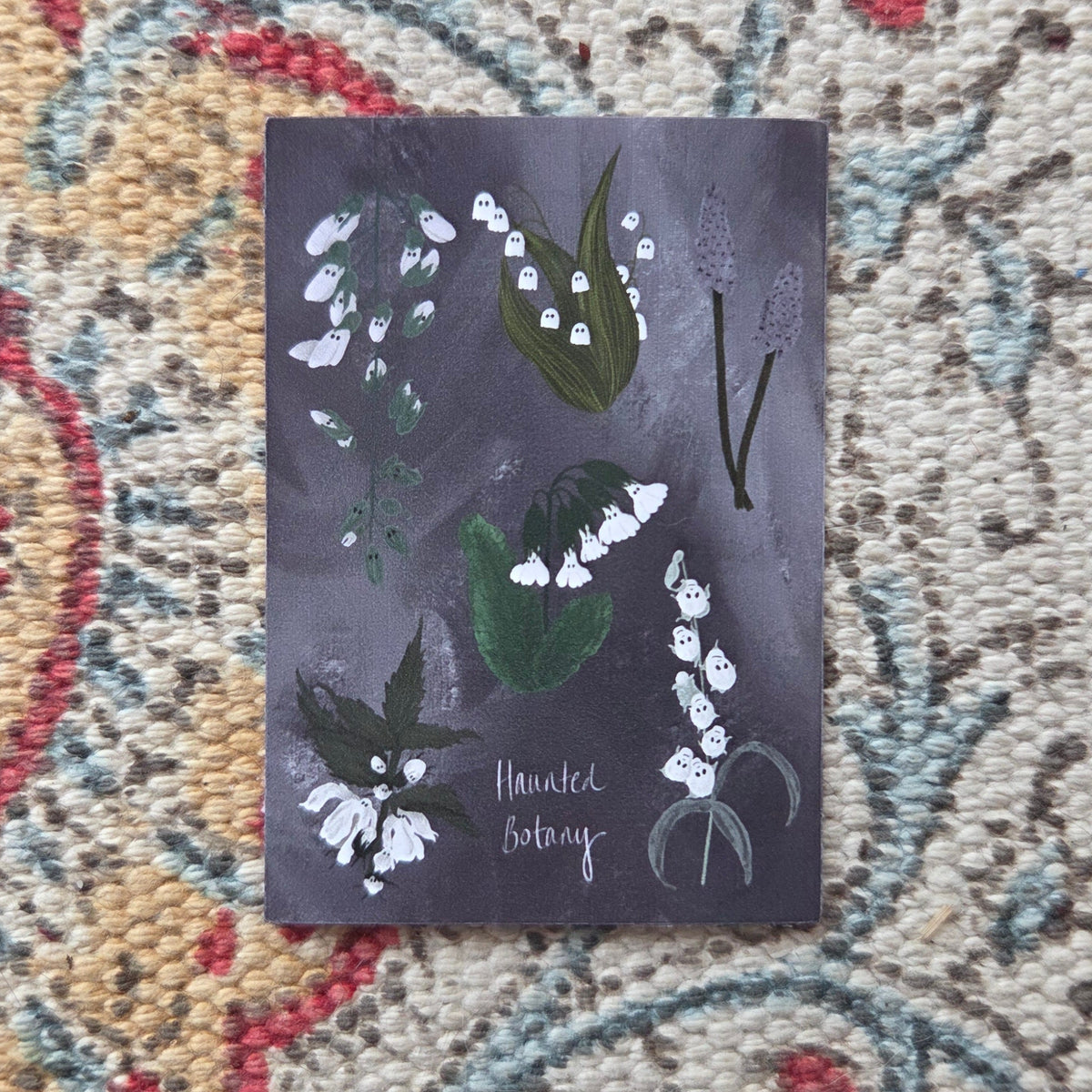 A purple and white rectangular sticker with six different haunted plant in whites and greens and the words 'Haunted Botany' at the bottom. Behind the sticker is a colourful patterned fabric.