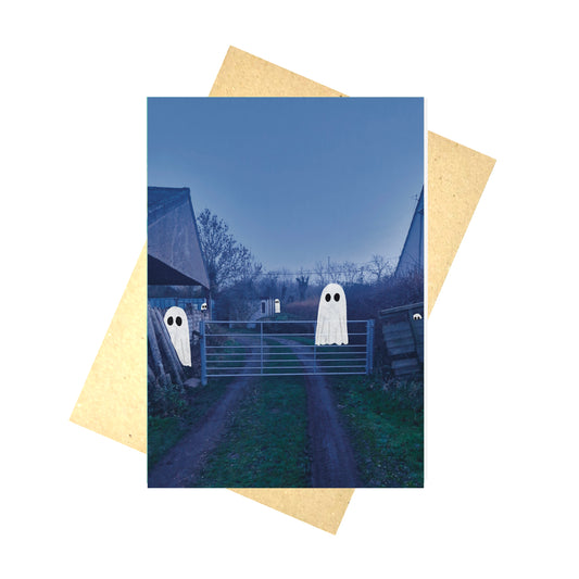 A blue card featuring a photo of a farm gate and track/barn with an assortment of ghosts. Behind the card is a brown paper envelope and a white background.