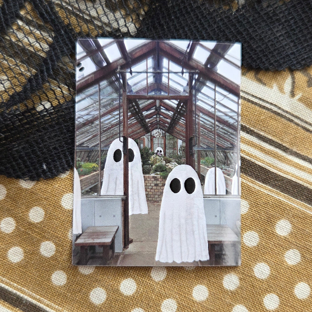 A small sticker featuring the Cambridge Botanics greenhouse with ghosts in it. The sticker is in front of a brown patterned fabric with a black lace overlay.