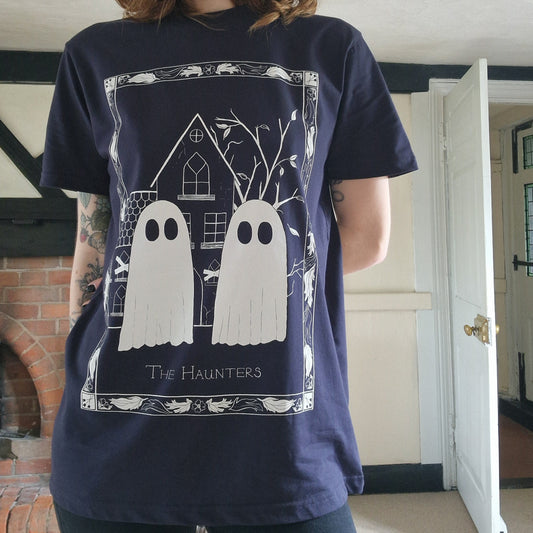 A tattooed person wearing a dark navy shirt with a white ghost design. Two ghosts float in front of an abandoned house within a patterned flower and leaf border. The person is also wearing black jeans and is standing in a white room with an old fireplace behind them and exposed black wood beams.
