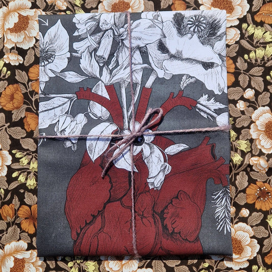 A dark grey tea towel sits folded on a retro floral background. The teatowel is tied with a purple twine and had a rich red heart illustration visible alongside some white poison plants illustrations.