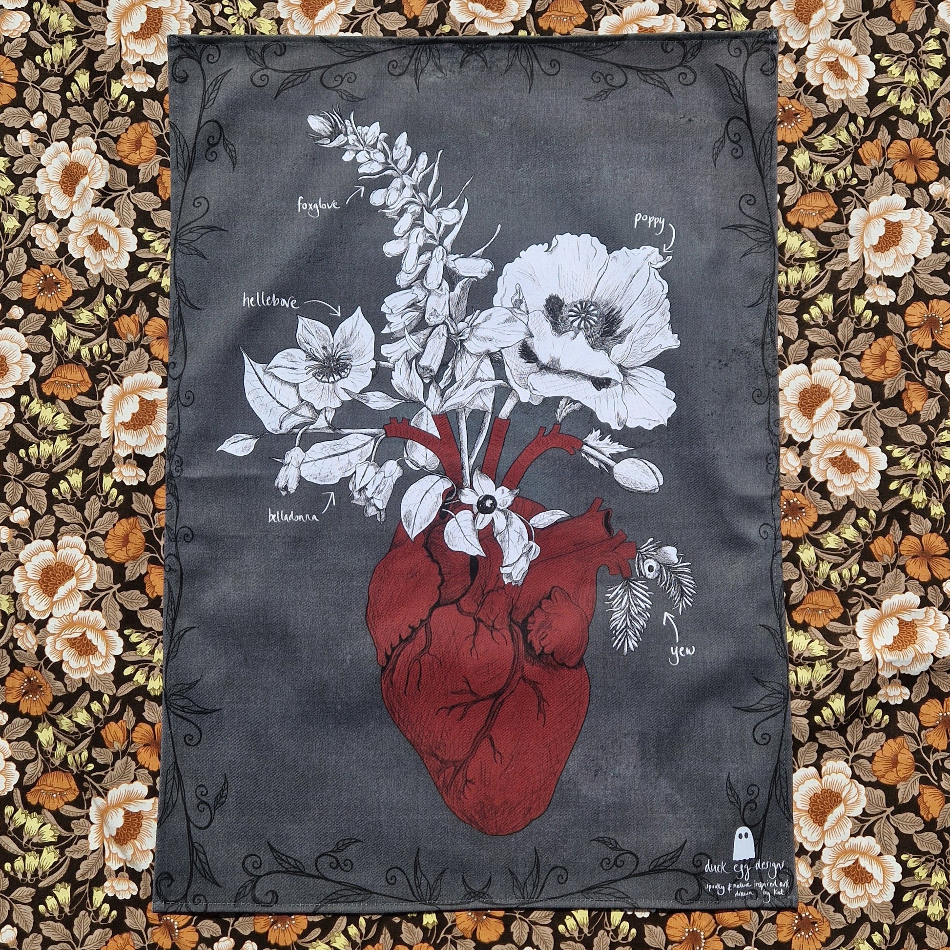 A dark grey tea towel lies on a retro floral patterned fabric. The teatowel has a anatomical red heart illustration with white and black poison plants growing from it. There's a black leafy border around the edges of the tea towel and a white Duck Egg Designs logo in the bottom right corner.