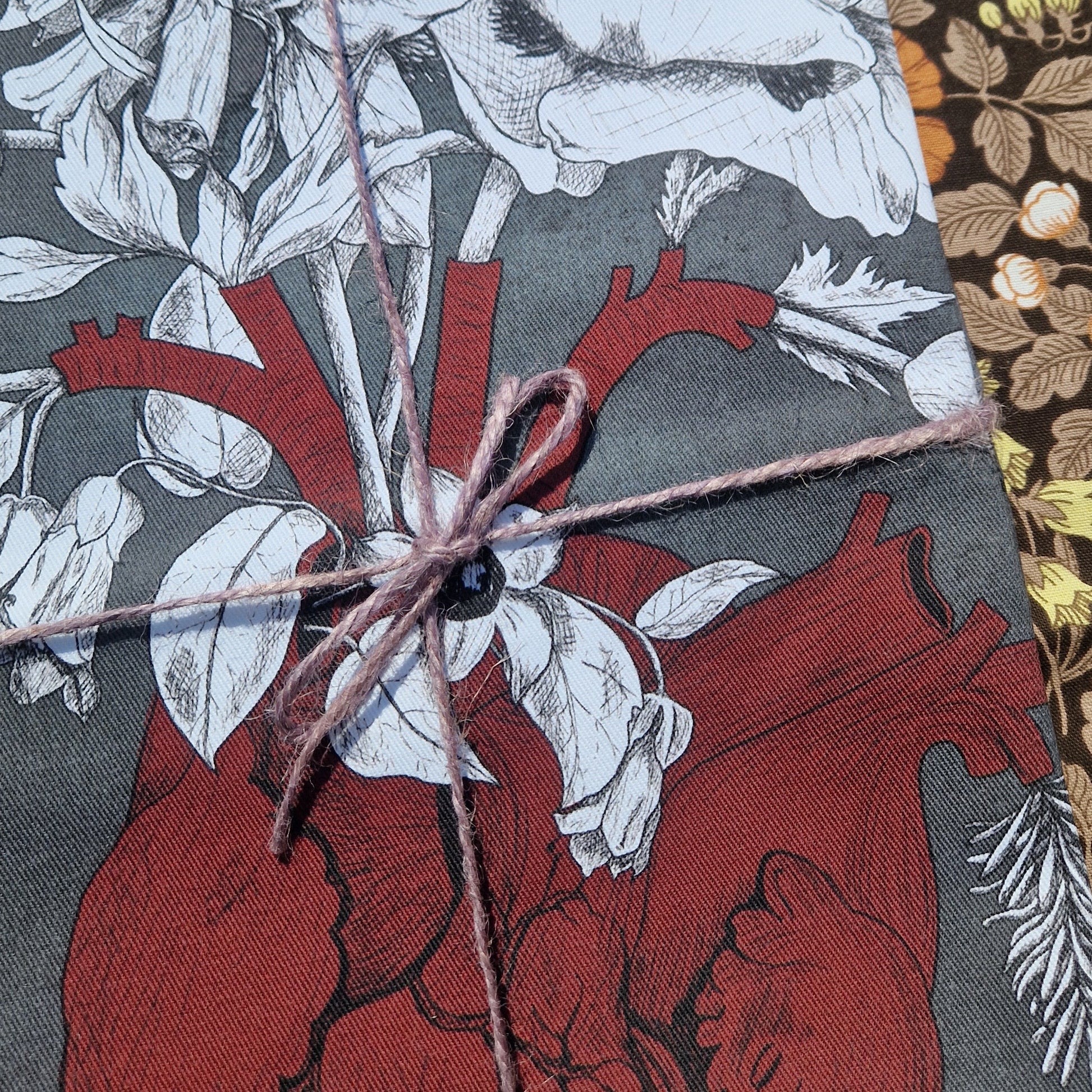 A close up view of the folded grey teatowel showing the red anatomical heart illustration and the white poison plants illustrations. The teatowel is wrapped with a purple twine and lies on a retro floral patterned background.
