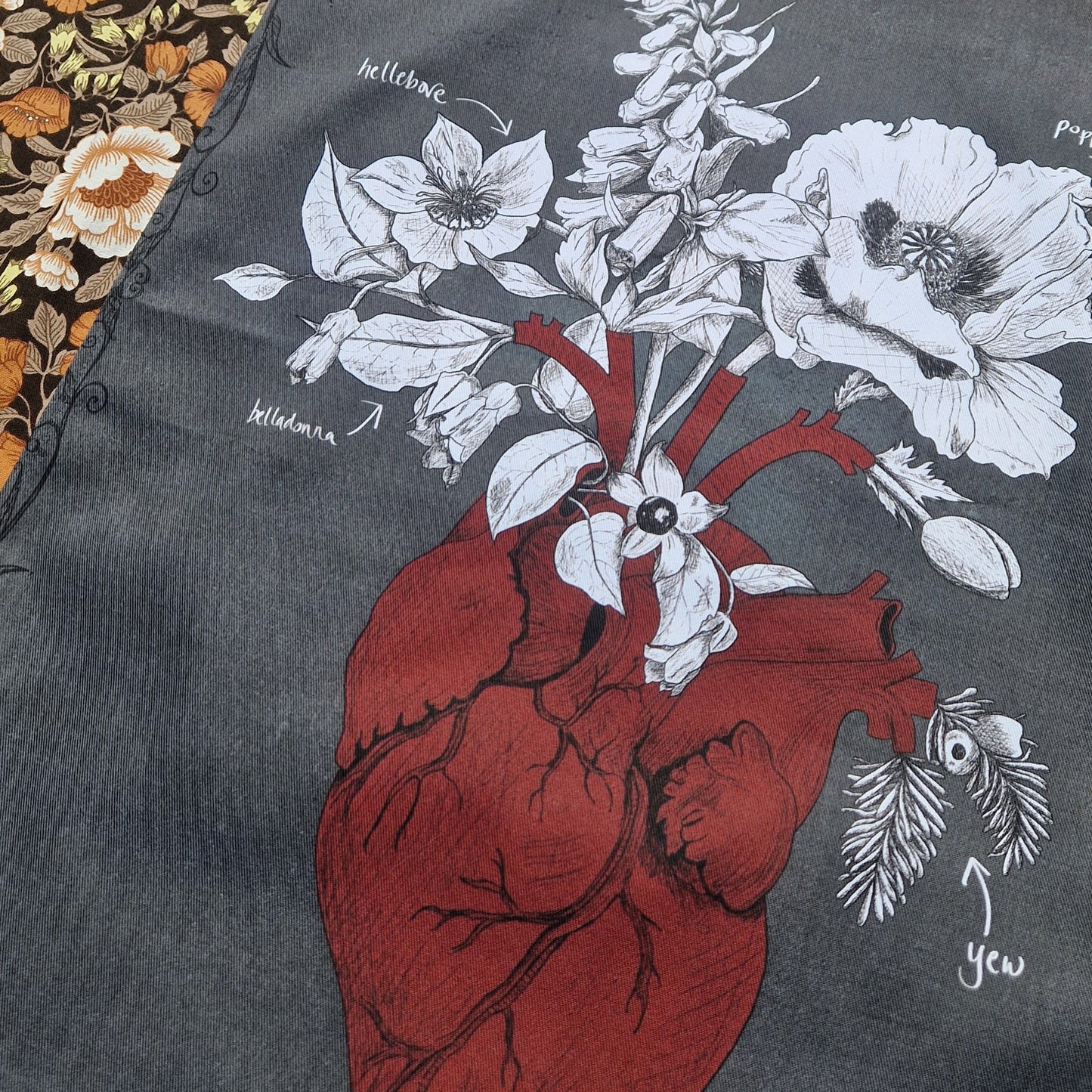 A close up view of the dark grey teatowel showing the rich red anatomical heart illustration and the white and black poison plants illustrations. 