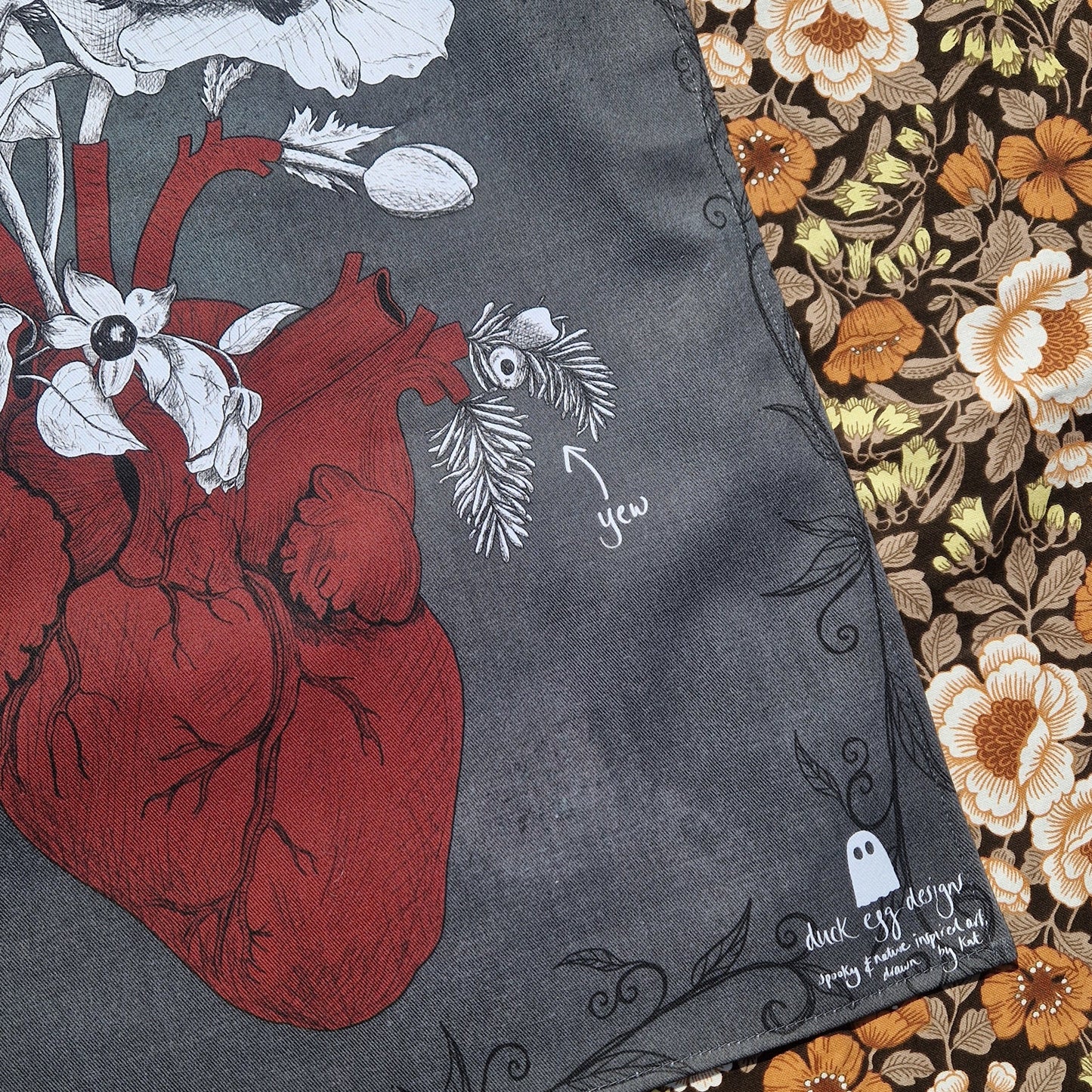 Close up of the bottom right of the dark grey teatowel showing the red anatomical heart illustration and the white Duck Egg Designs logo. To the right of the teatowel is a retro floral patterned fabric.