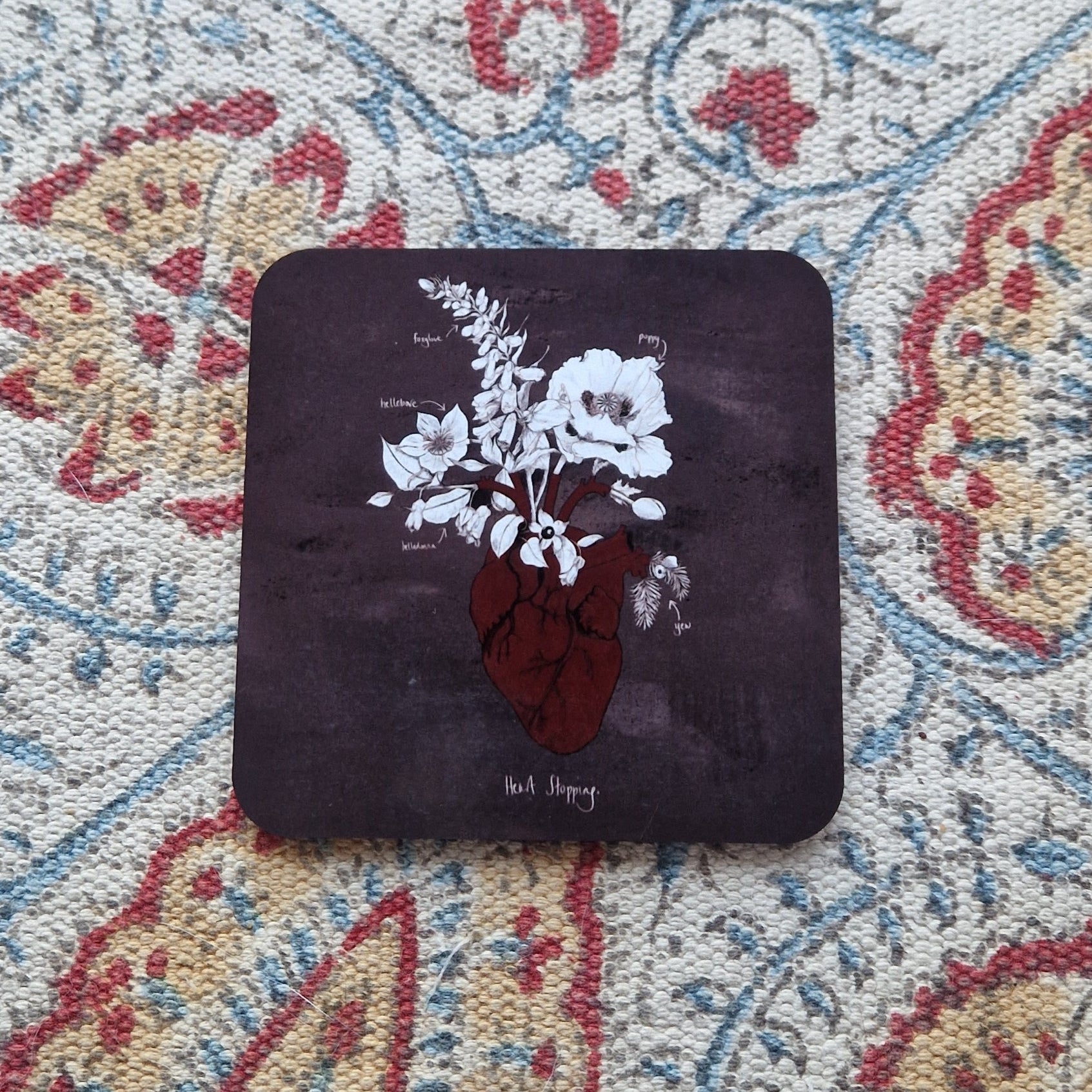 A mottled grey square coaster featuring a red anatomical heart illustration with white and black poison plants growing out from it. Behind the coaster is a patterned fabric background.