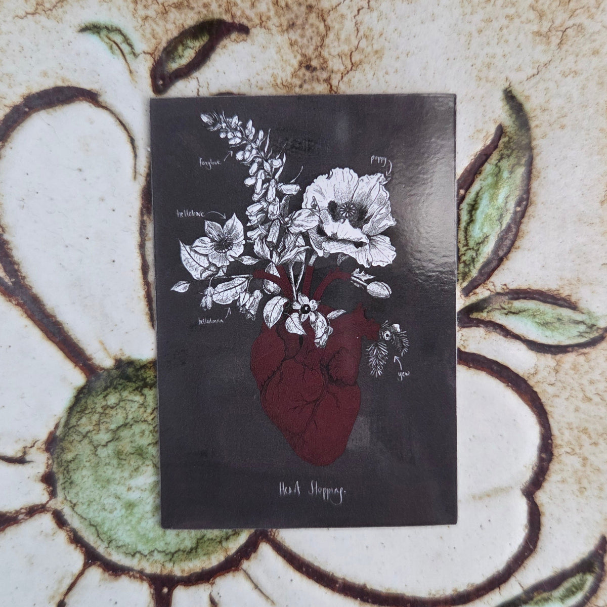 A grey rectangular sticker featuring a red anatomical heart with poison plants growing out of it and the words 'Heart Stopping' written in white underneath. Behind the sticker is a patterned tiled background.