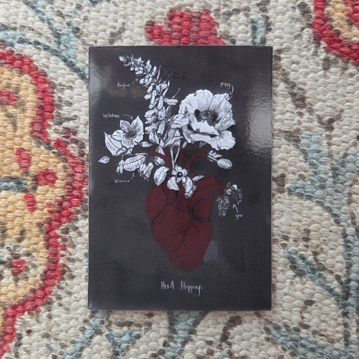 A grey rectangular sticker featuring a red anatomical heart with poison plants growing out of it and the words 'Heart Stopping' written in white underneath. Behind the sticker is a patterned fabric background.