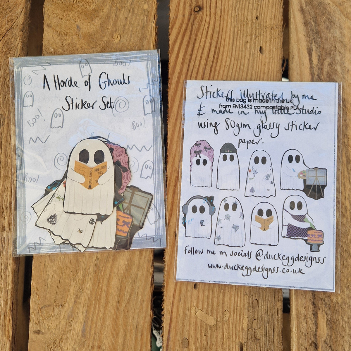 Two sticker sets lie side by side on a wooden plant background. The one on the left is top up showing the ghost stickers inside the pack with their black outlines. On the right the sticker set is face doing showing the information on the back and the eight different stickers in the set.