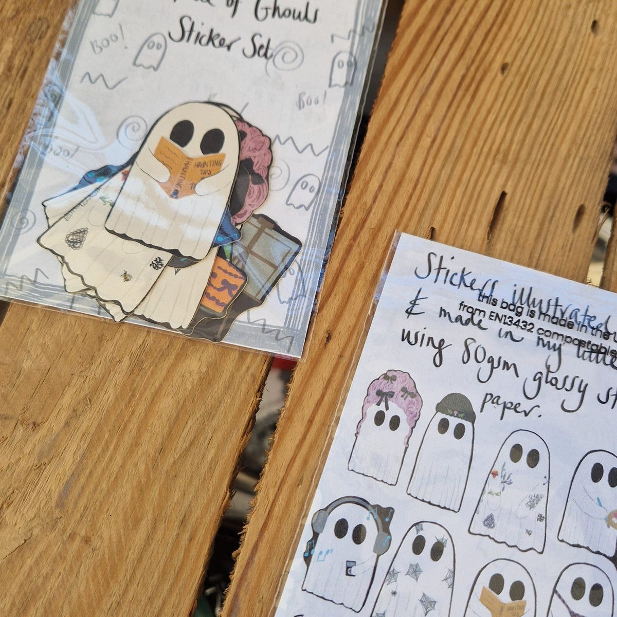 Two sticker sets lie side by side on a wooden plant background. The one on the left is top up showing the ghost stickers inside the pack with their black outlines. On the right the sticker set is face doing showing the information on the back and the eight different stickers in the set.