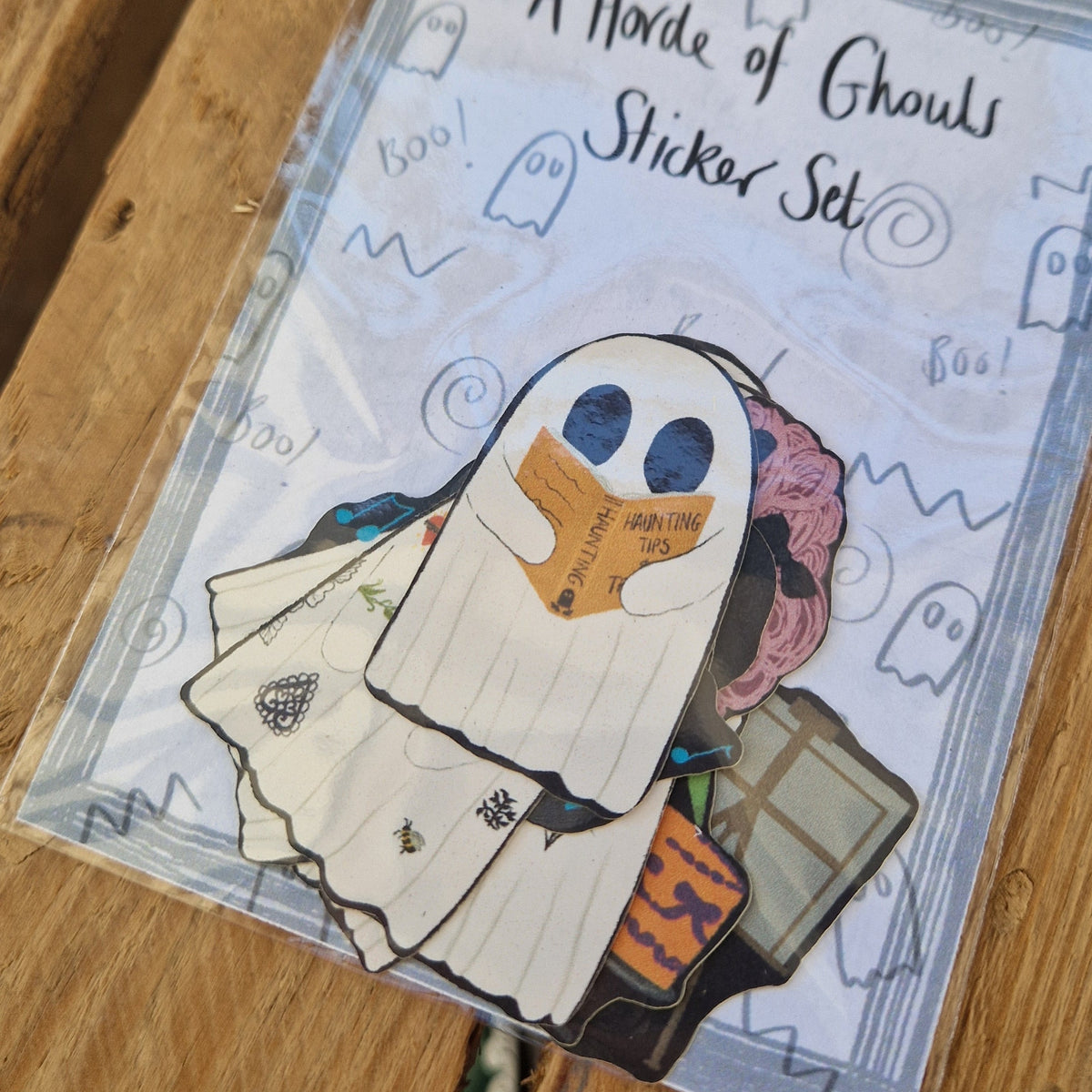 A close up of the front of the sticker set where you can see the eight ghost stickers inside - the sticker at the front is reading a book with an orange cover that reads 'HAUNTING TIPS'. Behind the sticker set is a wooden crate.