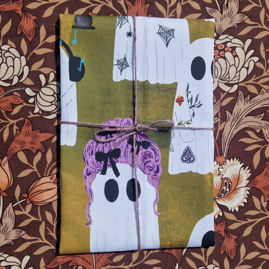 An orange folded tea towel with ghosts on it sits on a brown floral fabric. The tea towel has lilac twine wrapped around it and a ghost wearing a pink wig on the front.
