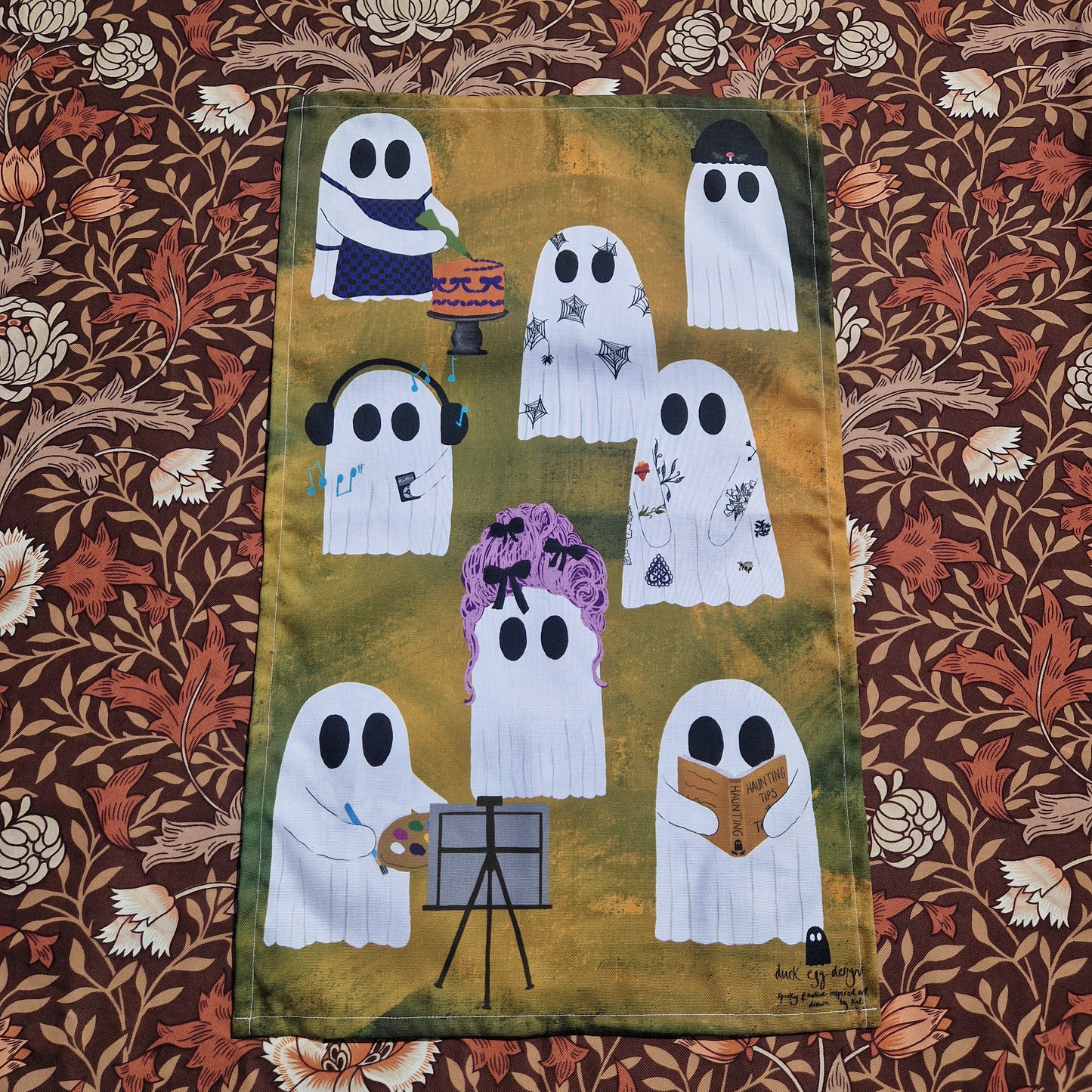 An orange and green tea towel with eight different ghosts sit on a brown floral patterned fabric. The ghosts all have different hobbies/outfits.