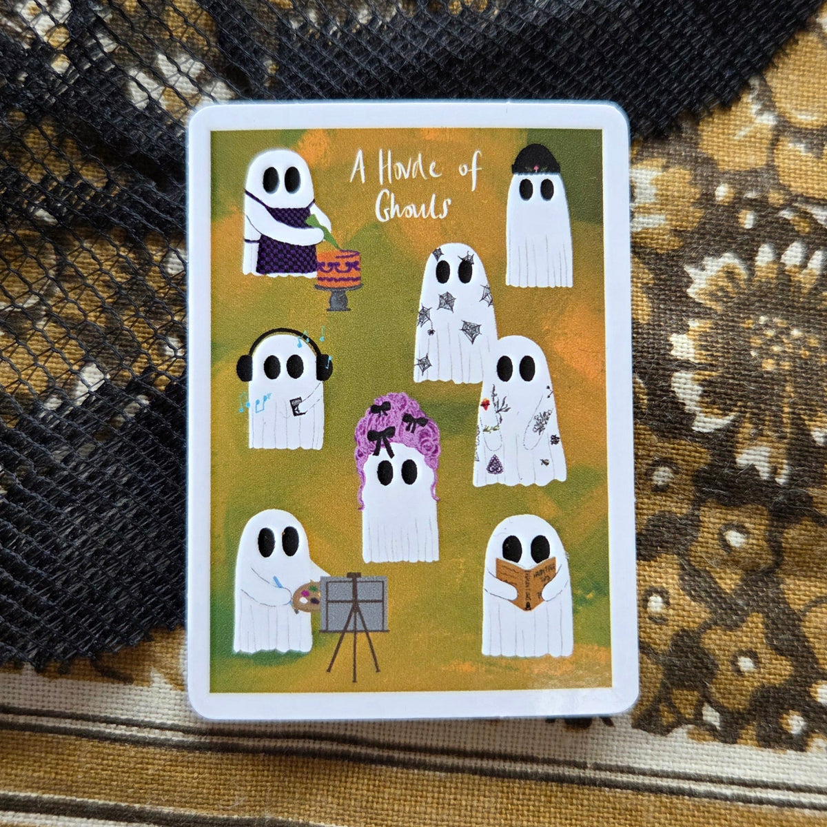 A rectangular orange sticker with a white border sits in front of a brown floral patterned fabric. The sticker features eight different ghosts along with the caption 'A Horde of Ghouls'.