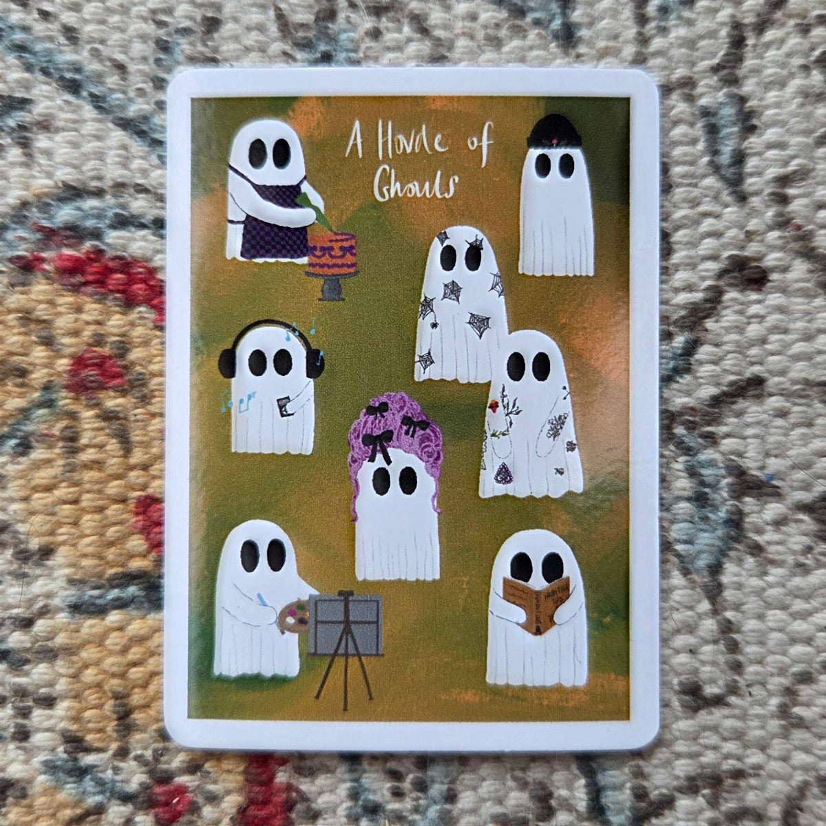 A rectangular orange sticker with a white border sits in front of a patterned fabric. The sticker features eight different ghosts along with the caption 'A Horde of Ghouls'. 